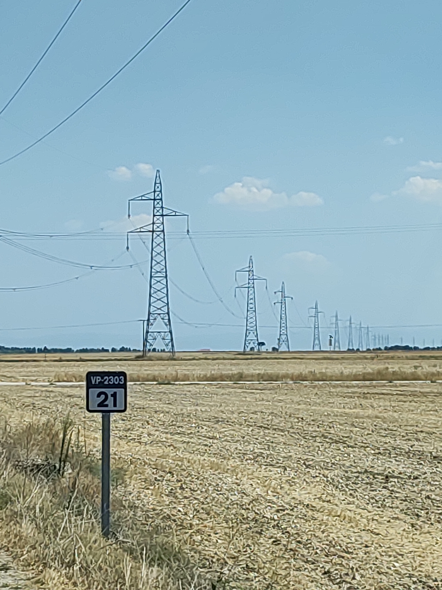 The image depicts a rural landscape in Cogeces del Monte, Spain, under a clear blue sky with scattered clouds. In the foreground, a field of dry, harvested crops stretches towards the horizon. A black and white signpost with the text "VP-2303" and the number "21" is positioned on the left side of the frame. A series of tall electricity pylons with high-voltage power lines traverse the scene from left to right, diminishing in size and clarity as they recede into the distance. The pylons are arranged in a linear fashion across the agricultural land. In the far distance, a treeline is visible, with a few buildings potentially present. The overall environment appears dry and open, characteristic of an agricultural area.