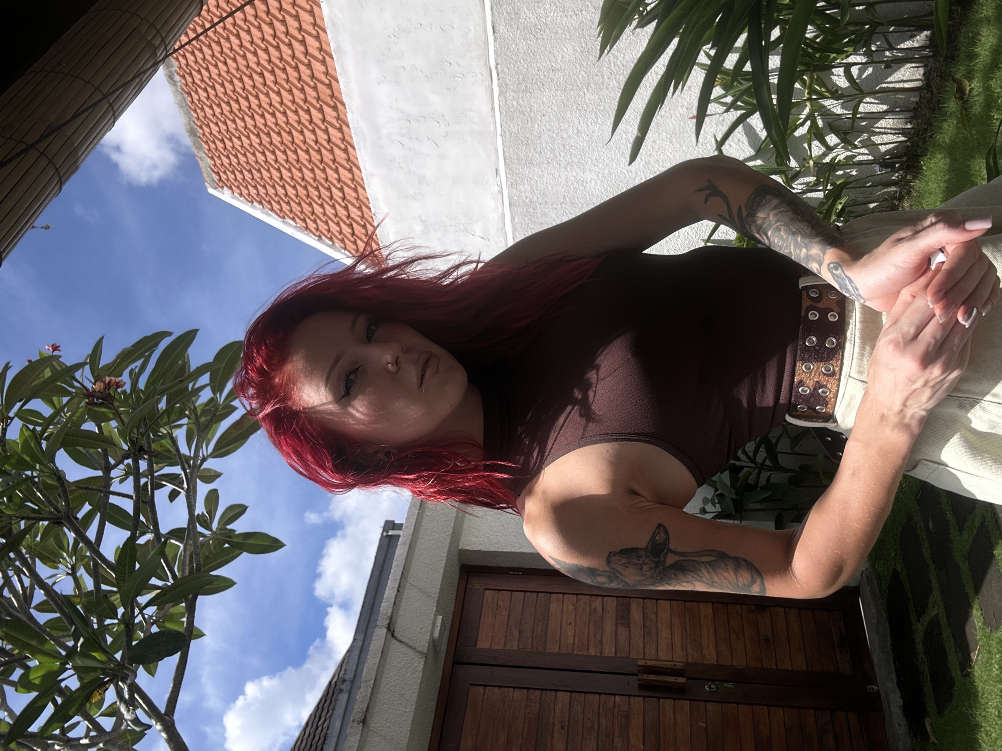 A young woman with long, vibrant red hair poses outdoors in North Kuta, Indonesia. She looks directly at the camera with a neutral expression, her hands clasped in front of her. She wears a dark brown sleeveless top and light-colored pants, accented by a brown belt with metallic eyelets. Her arms are adorned with extensive tattoos, including an animal design on her right bicep.
The setting appears to be a sunlit garden or courtyard. The background features a clear blue sky with scattered white clouds, a traditional red-tiled roof, and light-colored stucco walls. Lush green foliage from tropical plants, including one with small flowers, frames various parts of the image. A dark wooden door or gate is visible in the lower section, and patches of green grass are also present. Strong sunlight casts distinct shadows, indicating midday or early afternoon. No visible text is present.