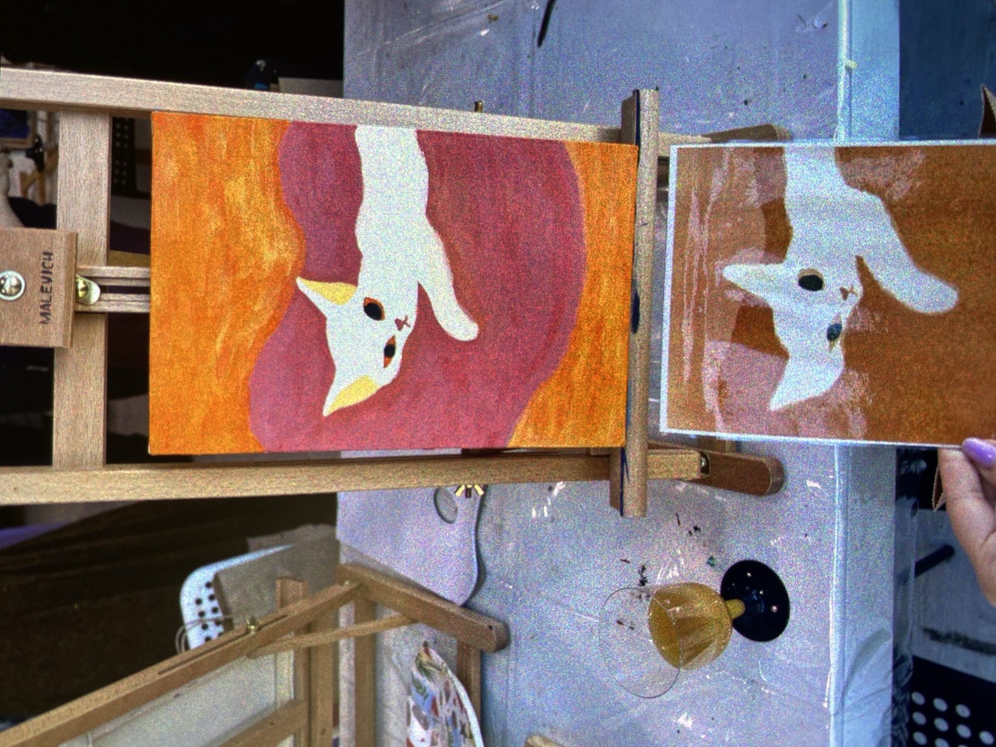 The image shows a painting on an easel featuring a stylized depiction of a white cat hanging upside down against an orange and pink background. Another similar, smaller painting is held beside it. The easel is made of wood and has the name "MALEVICH" printed on it. The paintings are rendered in a somewhat naive style. The background includes a partially visible table, another painting in progress, and various studio clutter. A hand with a light purple nail is holding the smaller painting. A transparent glass with a yellow substance is glued on the wall. The location is likely an artist's studio in Saint Petersburg, Russia, given the reference to Malevich, a prominent Russian artist, and the overall atmosphere of the scene.