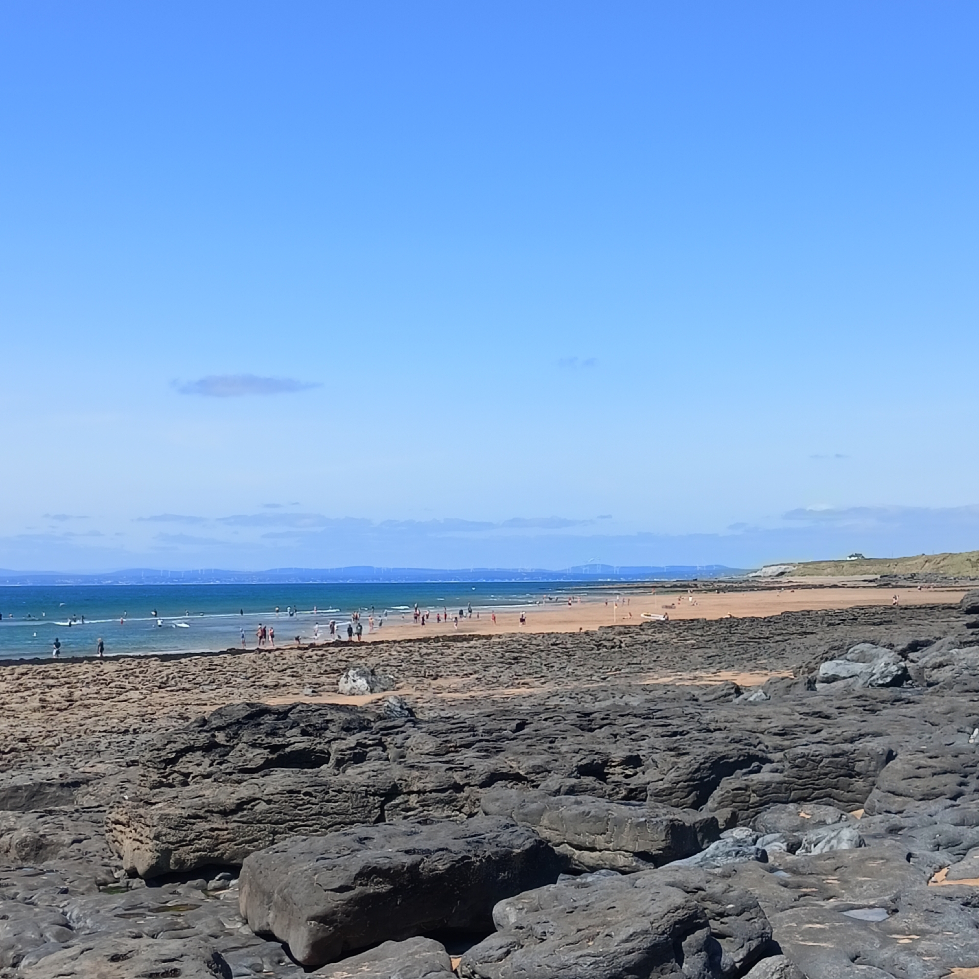 This image depicts a view of Fanore Beach in Fanore, Ireland, on a clear, sunny day. The foreground is dominated by large, dark, weathered rocks with uneven surfaces and some pools of water. Beyond the rocky shore, a sandy beach stretches out, populated by numerous people. Some individuals are walking along the shoreline, while others are in the shallow water, engaging in activities such as swimming or wading. Further out, the sea is a deep blue, with a line of distant land visible on the horizon, characterized by a range of hills dotted with wind turbines. The sky is a vibrant, cloudless blue.