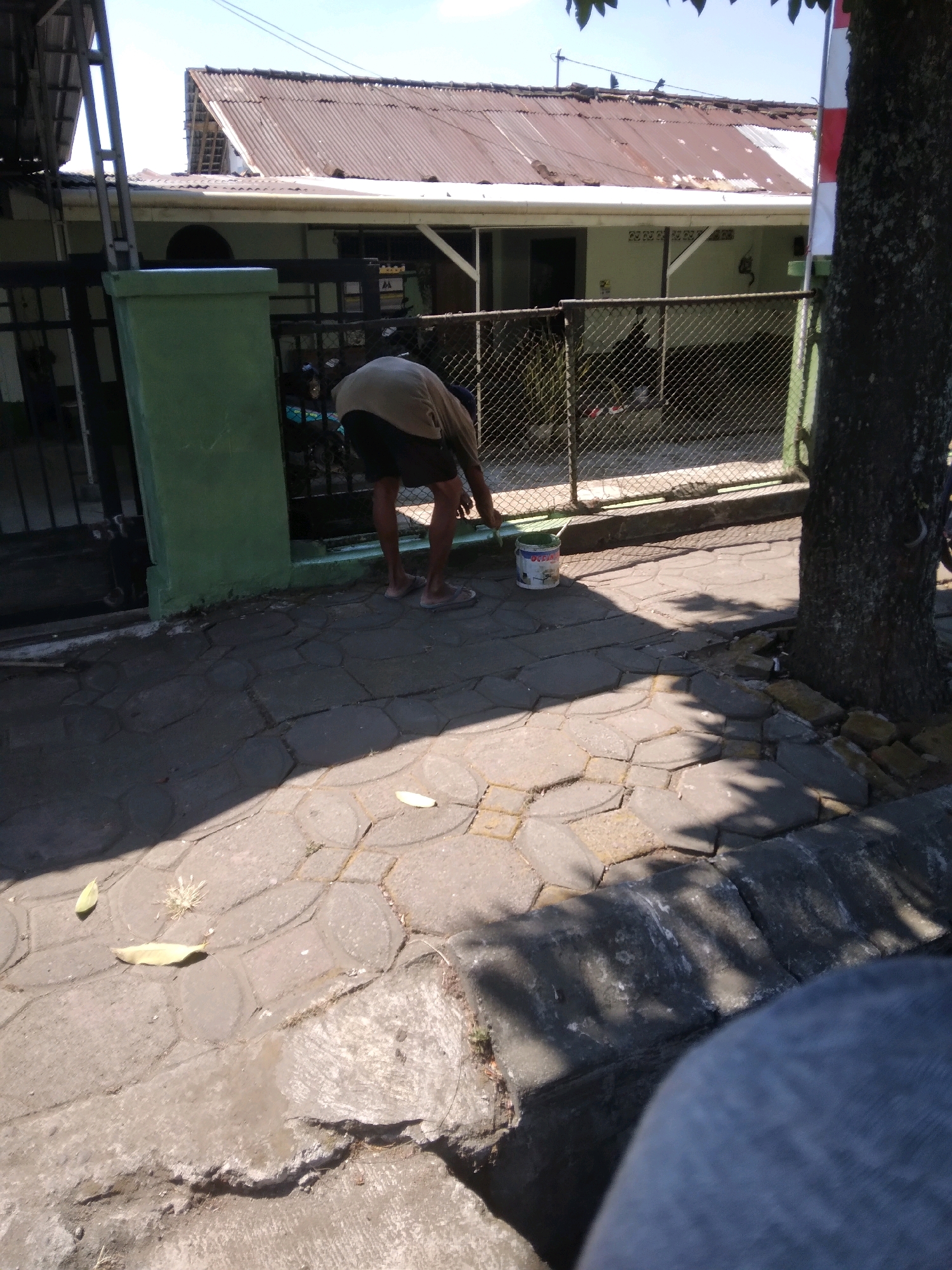 A person is bent over, painting a fence with a brush, standing in front of a building with a corrugated metal roof. A bucket of paint sits on the ground next to them. The scene appears to be outdoors, with a paved walkway and a tree. The location is Bancarkembar, Indonesia.