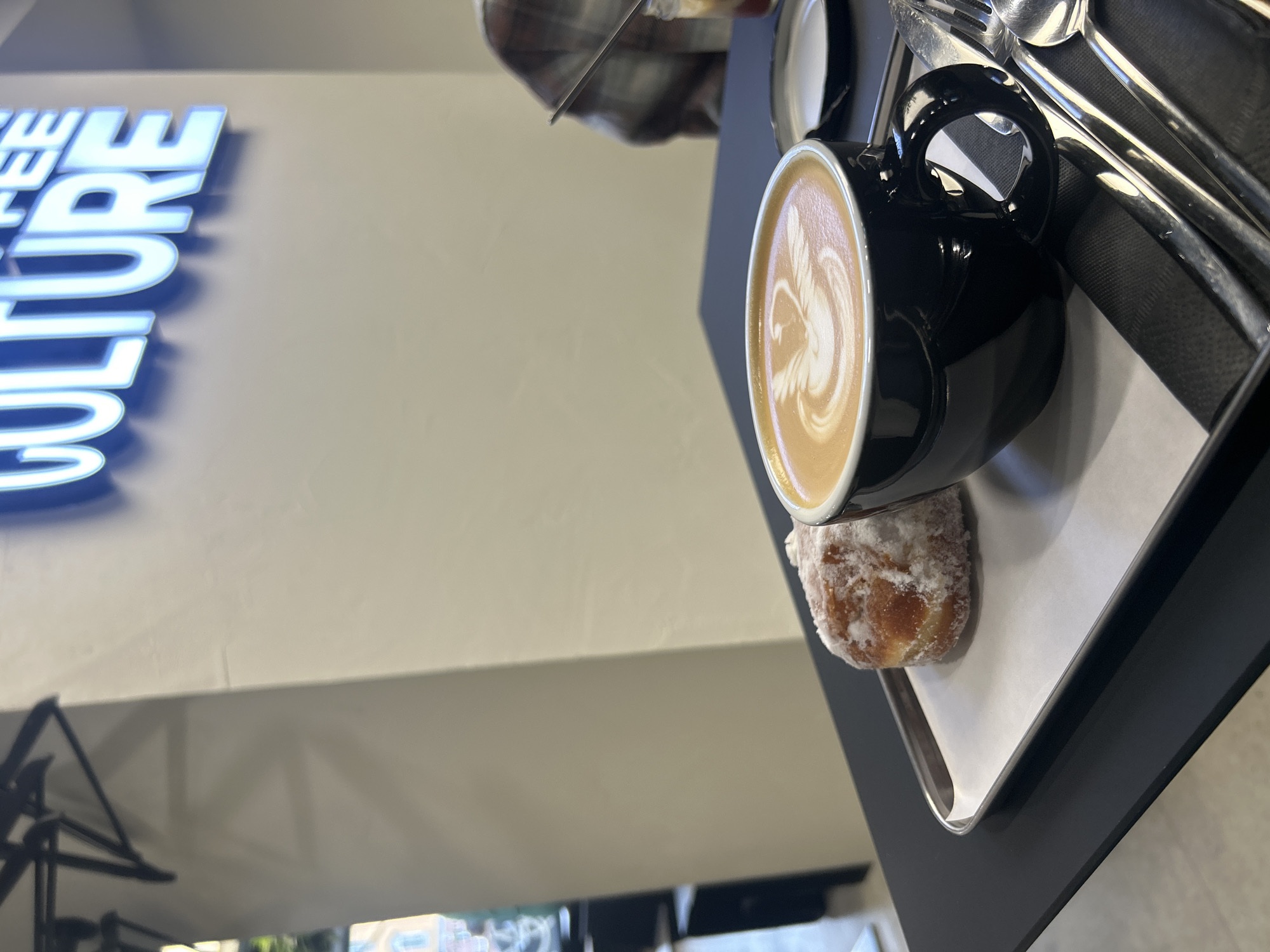 This image captures a close-up of a black ceramic mug filled with coffee, featuring latte art of a bird or swan. Beside the mug, on a white paper liner within a dark tray, rests a small, round pastry generously coated in powdered sugar. The scene is set in what appears to be a cafe, suggested by the "COFFEE CULTURE" sign with illuminated blue letters visible in the background. The lighting suggests it is daytime. A person, partially visible in the upper portion of the image, is wearing a plaid shirt and appears to be reaching for the cutlery placed in the tray. The overall impression is one of a relaxed moment enjoying coffee and a pastry.