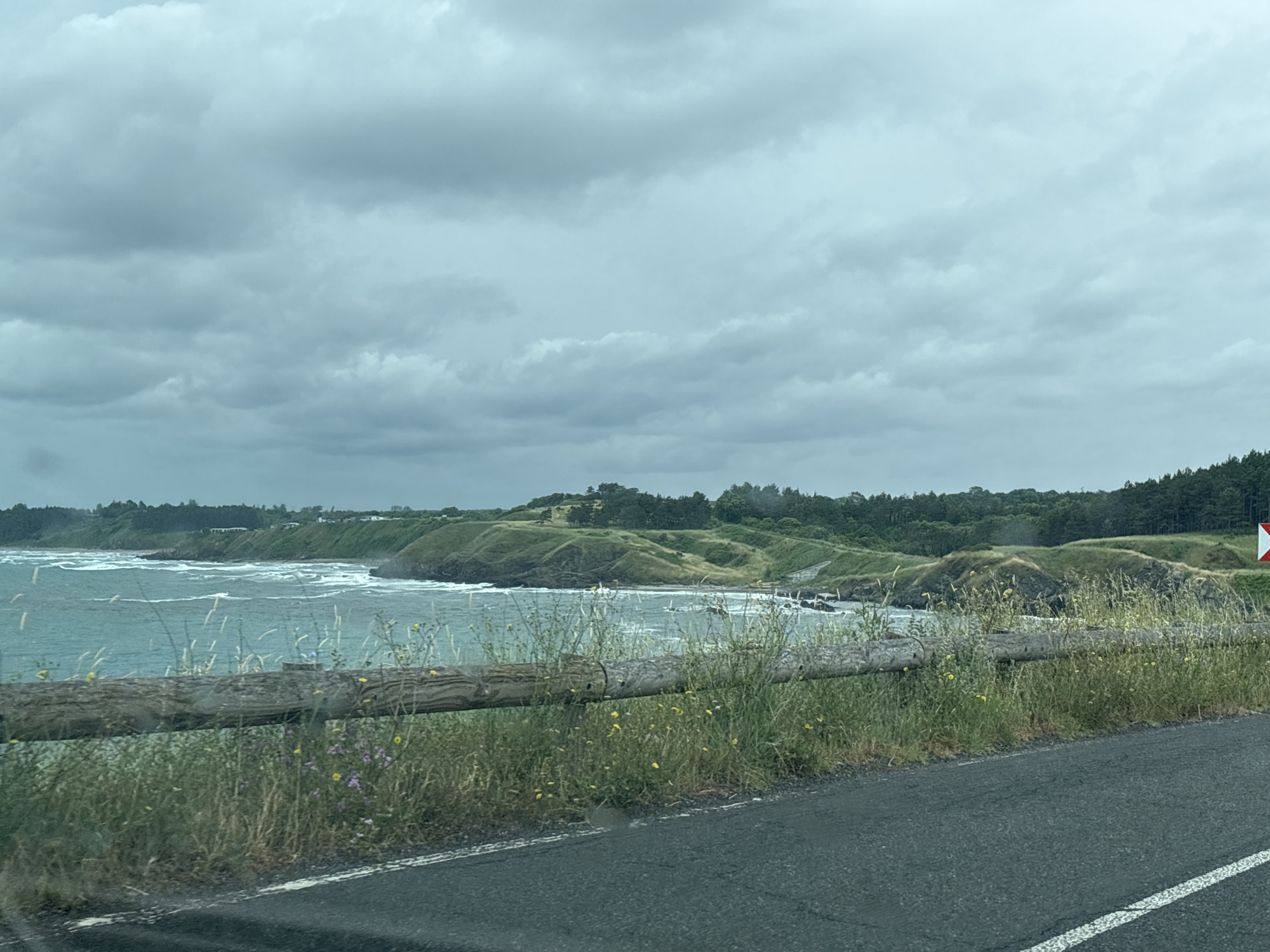 A cloudy day over the ocean. The water is a deep blue-green and choppy, with whitecaps. In the foreground, there is a wooden fence, and beyond it, green hills dotted with trees. The road is asphalt and runs along the coast. A white line is visible on the right side of the road. The scene appears to be a coastal highway, possibly in Bulgaria, given the location context.