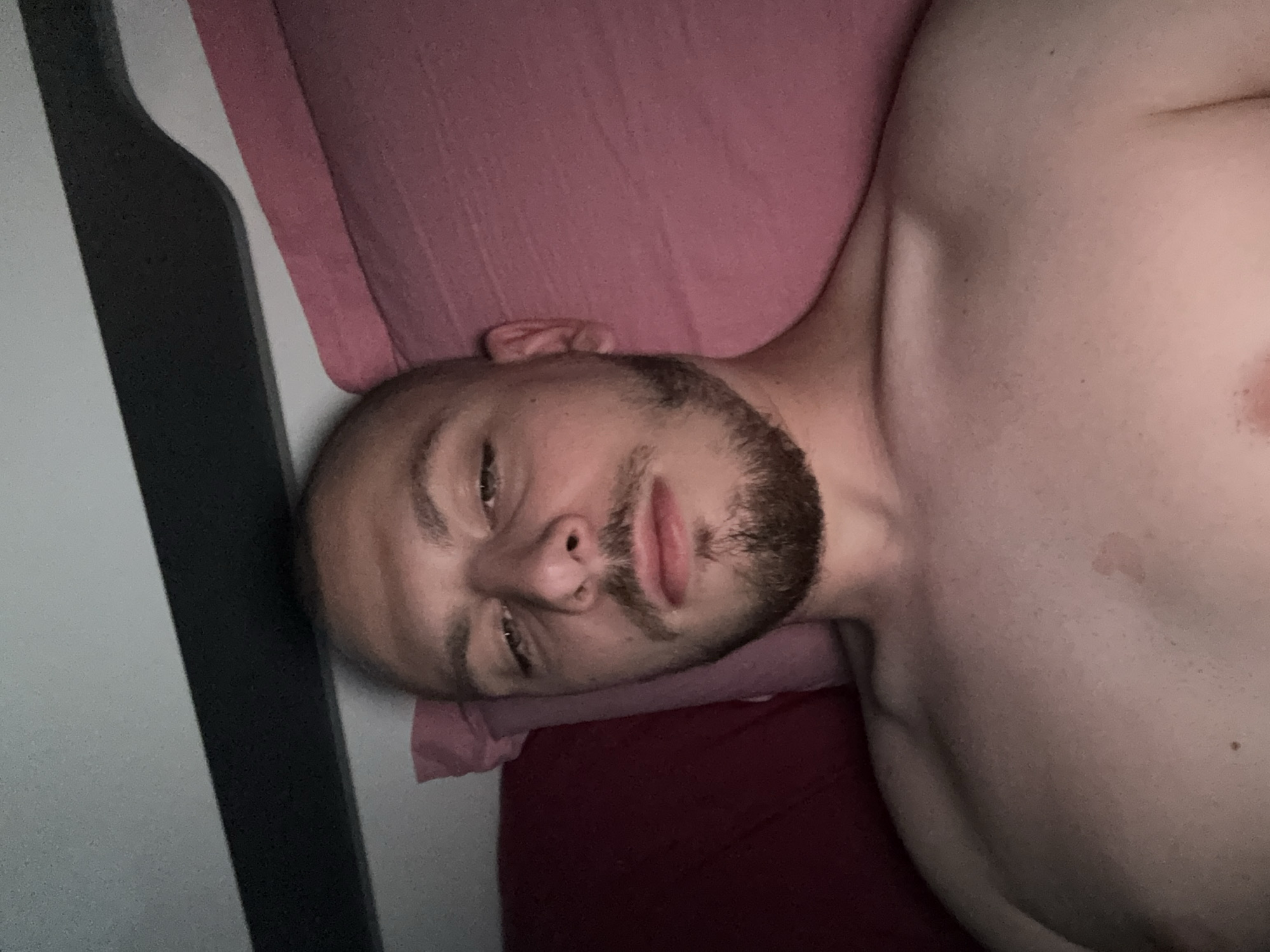 An adult male with fair skin, a shaved head or very short hair, and a trimmed beard, appears to be in his late 20s to early 30s. He is lying shirtless on a bed or similar surface, looking directly at the camera. His head rests on a light pink pillow, with a darker red pillow visible to his left. Pink bedding extends across the right side of the frame. Above his head, a dark horizontal structure, possibly a headboard or shelf, is visible against a lighter wall or ceiling. The lighting is dim, suggesting an indoor environment. The image was captured in Stara Zagora, Bulgaria.