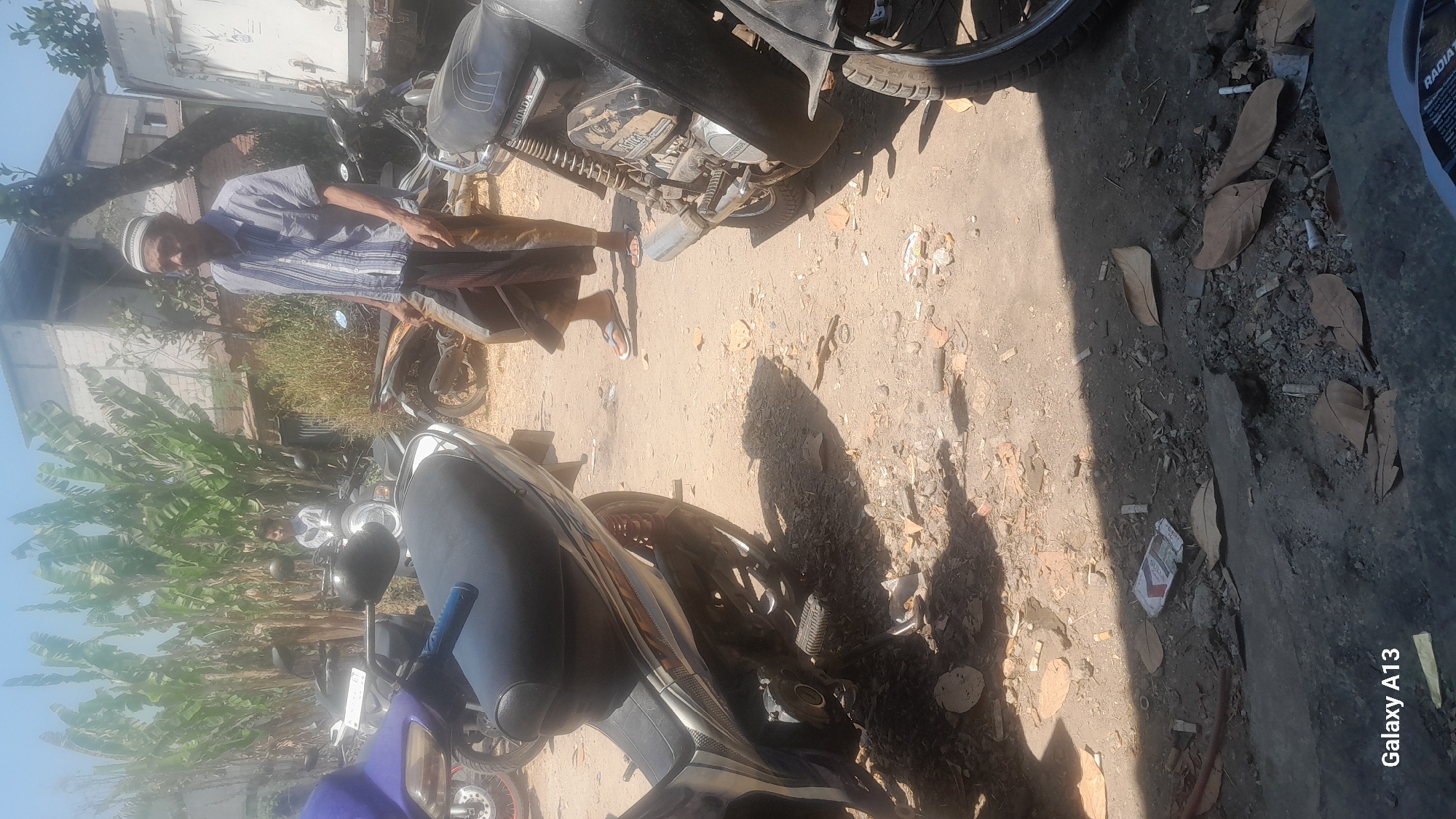 The image depicts an outdoor scene, likely in a city. In the foreground, a motorcycle is partially visible, with its seat and rear wheel in focus. The ground is dirt and gravel, with scattered leaves and cigarette butts. A person in the background, an elderly man wearing a striped shirt and a white skullcap, is standing near a motorcycle. He is wearing a sarong and sandals. There are several other motorcycles and a large white truck in the background. Banana trees are visible behind the people and vehicles. The overall impression is one of a casual gathering or a parking area, possibly in a warm, sunny climate. The presence of a "Galaxy A13" watermark in the bottom left corner indicates that the image was taken with a Samsung Galaxy A13 smartphone. The location is identified as Hadipolo, Indonesia.