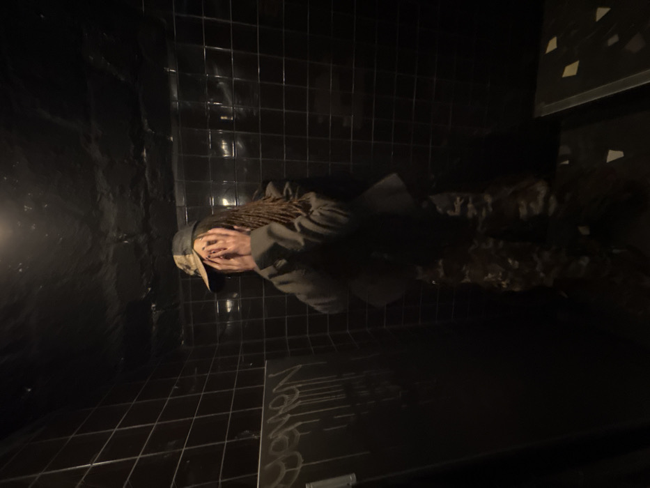 A person is crouched in the corner of a dark, tiled room. The person's face is obscured by their hands, and they are wearing a camouflage hat and a dark jacket. The room appears to be a bathroom, with dark tiles on the walls and floor. There are faint inscriptions on the floor. The lighting is dim and uneven, with shadows obscuring some of the details.
