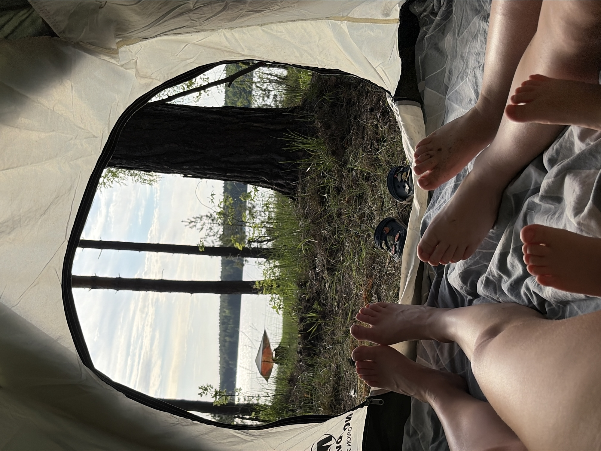The image, taken from inside a tent, shows a view of Забегаево, Russia. The foreground features several pairs of bare human legs and feet resting on a gray blanket inside the tent. Just outside the tent opening, the ground is covered with grass and plants. Beyond that, the landscape reveals a body of water surrounded by trees. A single orange kayak or paddleboard is visible in the water. The sky is overcast. Two pairs of Crocs sandals are visible near the legs.