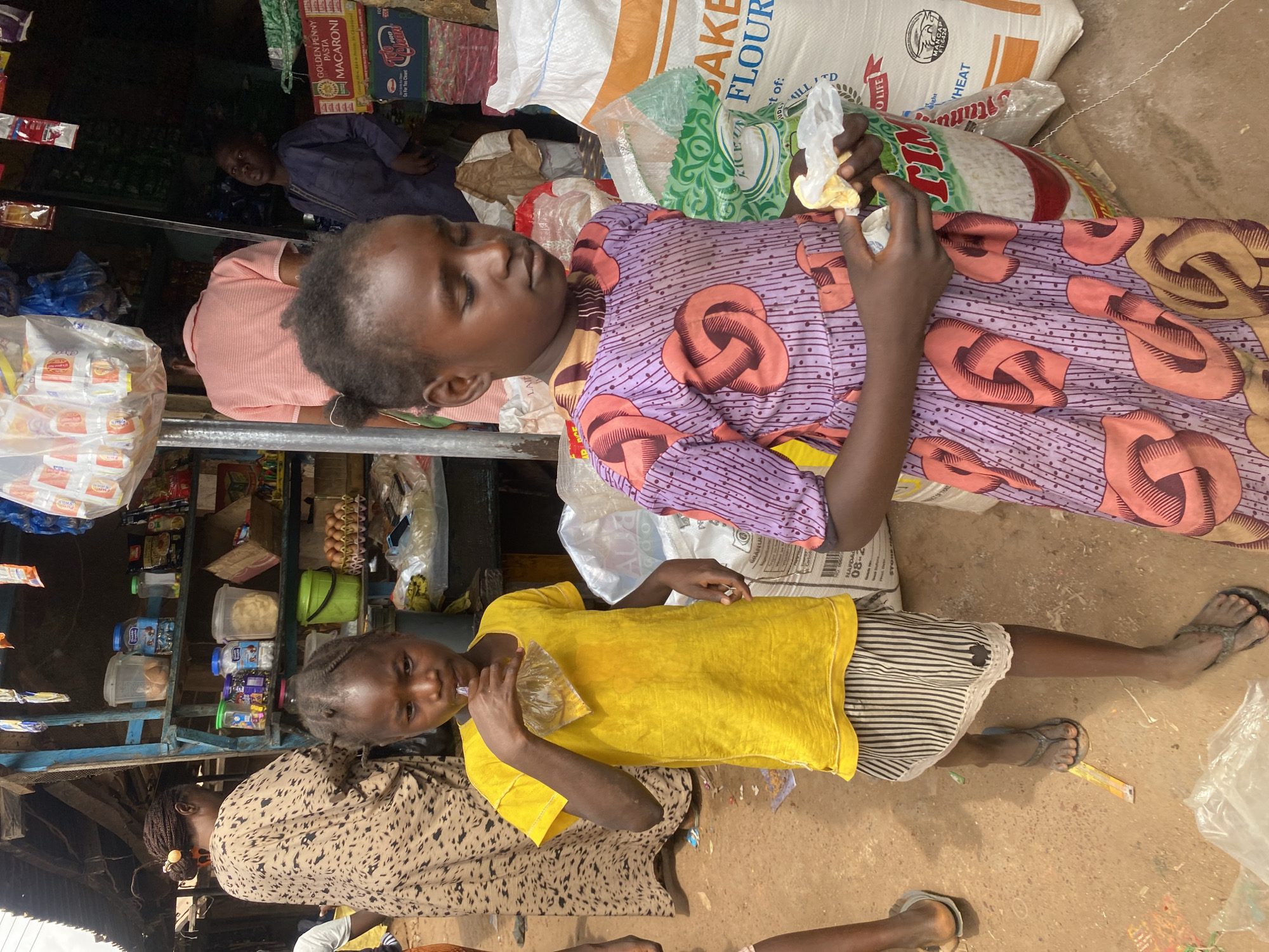 The image depicts a bustling outdoor market or roadside shop in Rido, Nigeria. In the foreground, two young girls, approximately 6-9 years old, are prominent. The girl on the left wears a bright yellow t-shirt over a black and white striped skirt and dark flip-flops; she is actively drinking from a transparent plastic sachet containing a yellowish liquid, holding it to her mouth with her right hand. Her left hand hangs at her side. The girl on the right, with braided hair, wears a purple dress adorned with an orange-brown repeating loop pattern. She is looking downwards, focused on a crumpled white wrapper, possibly containing food, held in her hands. She stands next to a large white sack labeled "BAKE FLOUR."
In the midground, to the left, a woman is seen from behind, wearing a loose-fitting, light-brown patterned dress, with a yellow hair tie visible in her braided hair. She appears to be facing shelves within the shop. Behind the girl in yellow, another person, seen from behind, wears a light pink short-sleeved shirt. To the right, further back, a boy or young man in a dark blue, long-sleeved garment looks upwards and to his left.
The shop structure features blue wooden posts supporting a corrugated metal roof. Shelves behind the figures are stocked with various goods: clear plastic containers hold snacks like biscuits or pastries, along with bottles and smaller packaged items. Multiple cardboard trays of eggs are visible on a lower shelf. Large sacks of provisions, including "GOLDEN PENNY PASTA MACARONI" and "PRO CEREAL," are stacked in the background. Numerous small packets and sachets of unknown contents hang from lines above the shelves, and a large clear plastic bag filled with white packets is suspended overhead. The ground is unpaved, reddish-brown dirt, with scattered litter, including discarded plastic. The scene is illuminated by natural daylight, indicating an outdoor or semi-open setting.