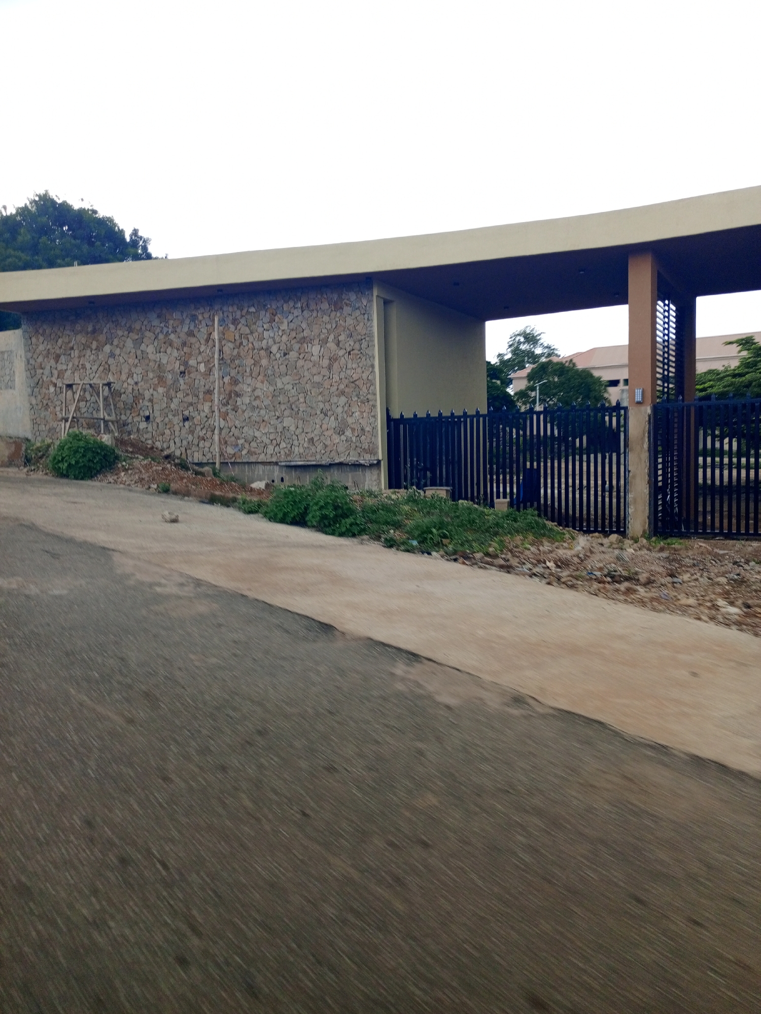 The image shows a modern building with a curved roofline, constructed with stone and concrete. A black metal fence with vertical bars encloses the property. A paved pathway leads up to the fence and into the building's entrance, which is partially visible. Green vegetation and some scattered debris are present on the ground. The sky is overcast. The scene appears to be an exterior view of a commercial or institutional building.