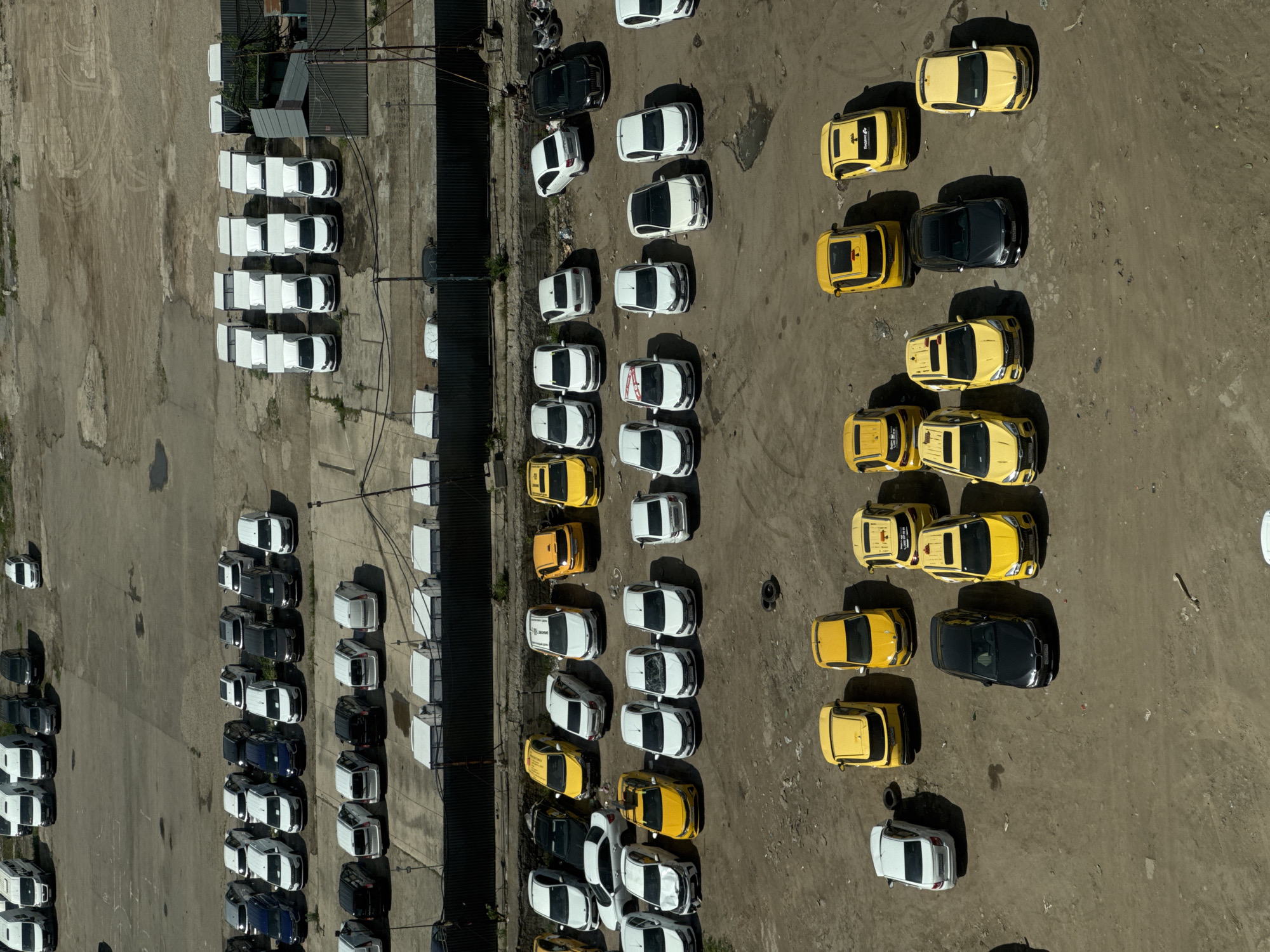 The image shows an aerial view of a parking lot in Ukromne, Ukraine. The lot is filled with numerous cars, primarily arranged in orderly rows. The cars are a mix of colors, including white, black, yellow, and blue. The surface of the parking lot is predominantly concrete. To the left side of the frame, there is a structure appearing to be a building.