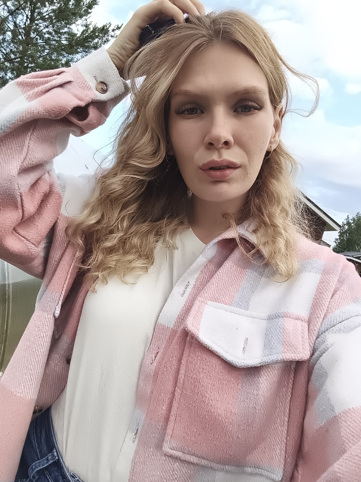 A young woman with blonde, wavy hair is pictured in a close-up selfie. She is wearing a pink and white plaid shirt with a white t-shirt underneath, and blue denim jeans. Her right hand is raised and touching her hair. Her expression is neutral, and she is looking directly at the camera. The background shows a cloudy sky, a green tree, and the roof of a building. The overall lighting suggests it is daytime, likely afternoon, with soft, diffused light. No text is visible in the image. The scene appears to be outdoors in a rural or suburban setting, possibly in Русский Вожой, Russia, given the location context.