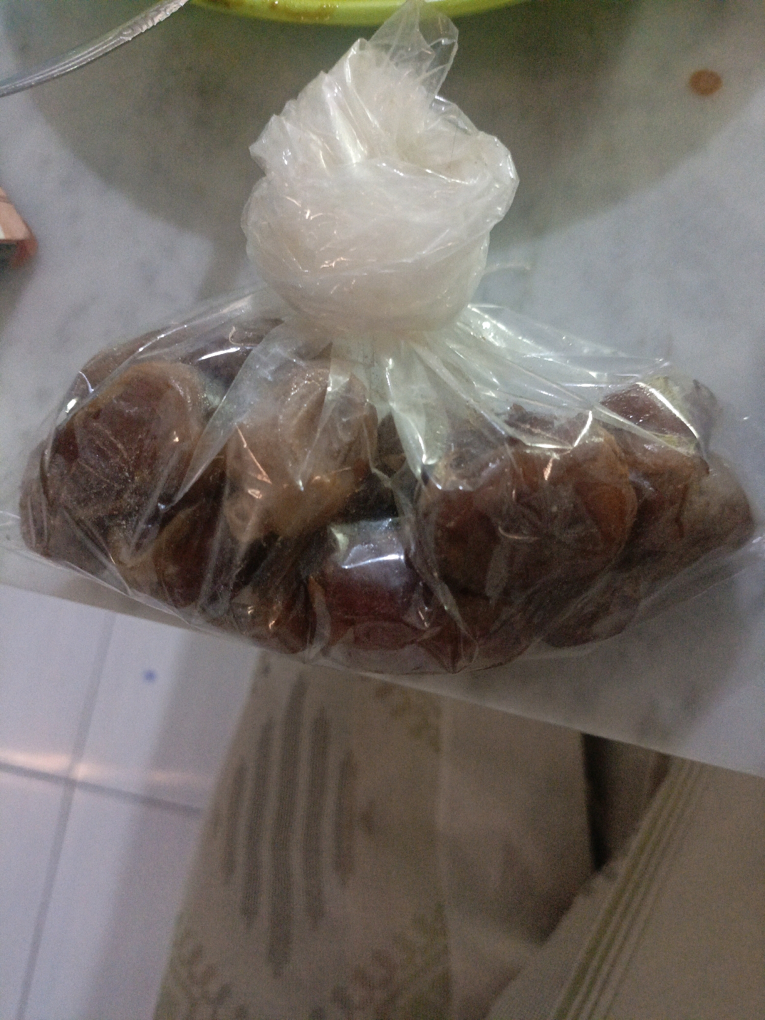 A plastic bag filled with dates is placed on a light-colored surface. The bag is translucent, and the dates inside are dark brown and appear to be whole. Some of the dates have a powdery or crystallized coating. The bag is tied at the top with a twisted piece of plastic. The background is out of focus and appears to be a neutral, possibly indoor environment. There is a hint of a green object in the upper left corner of the image, possibly a bowl or plate. The lighting in the image is soft, with shadows cast by the bag and its contents. The scene is devoid of any people or animals. The location is Undaan, Indonesia.