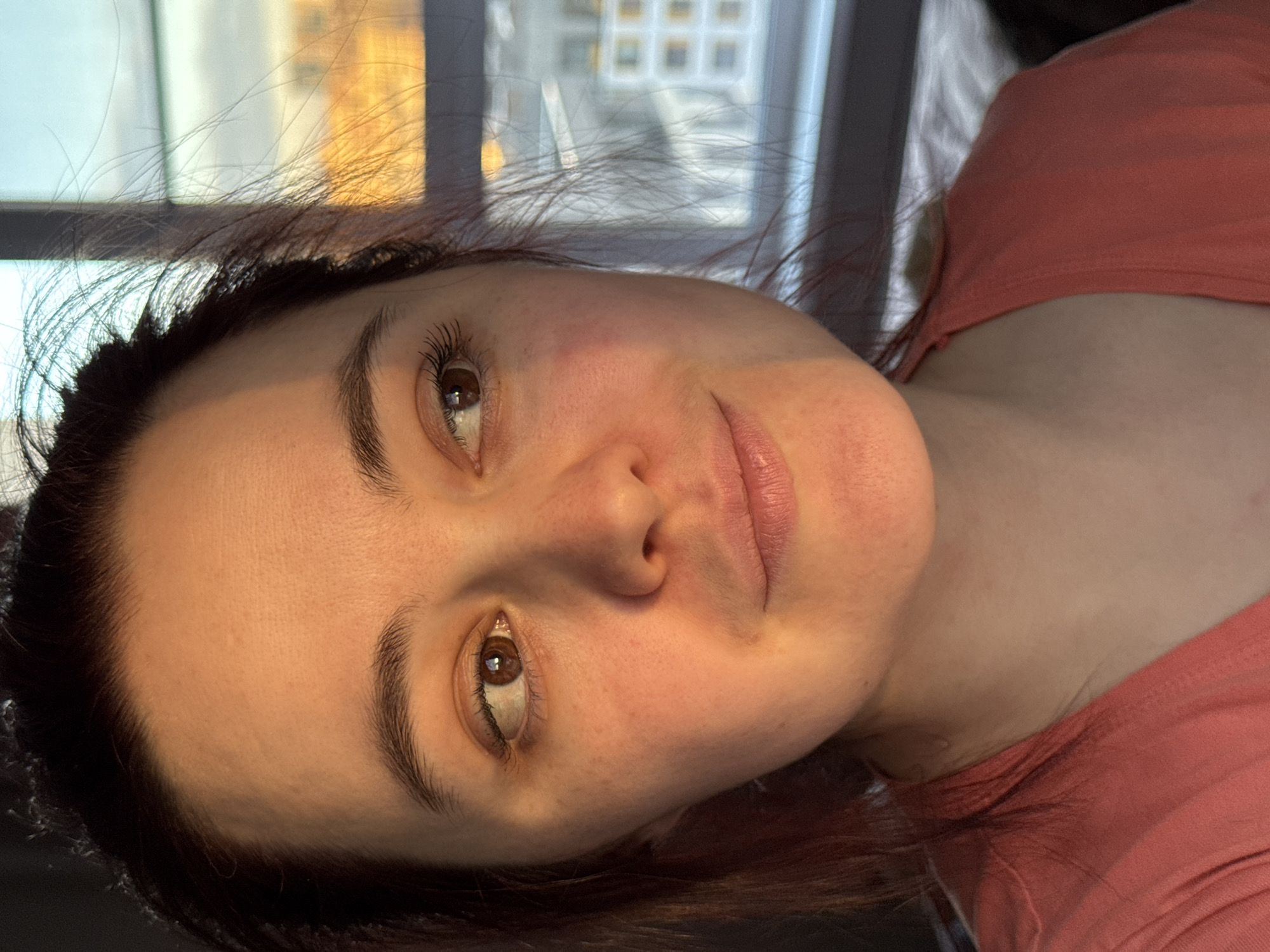 A close-up, eye-level portrait of a woman is shown, with her head tilted to the left, looking directly at the viewer. She has warm brown eyes, dark brown hair that is slightly disheveled, and light skin, with a natural, subtle expression hinting at a slight smile. Her eyelashes appear to have mascara. Strong, warm, golden light, likely from the sun, illuminates the right side of her face and hair, creating highlights. She is wearing a reddish-pink top. In the background, visible through a window, is a multi-story building with a grid of windows, indicating an urban setting. The lighting suggests it is late afternoon or early evening. The woman appears to be reclining or lying down indoors, possibly in Izhevsk, Russia. No text is visible.