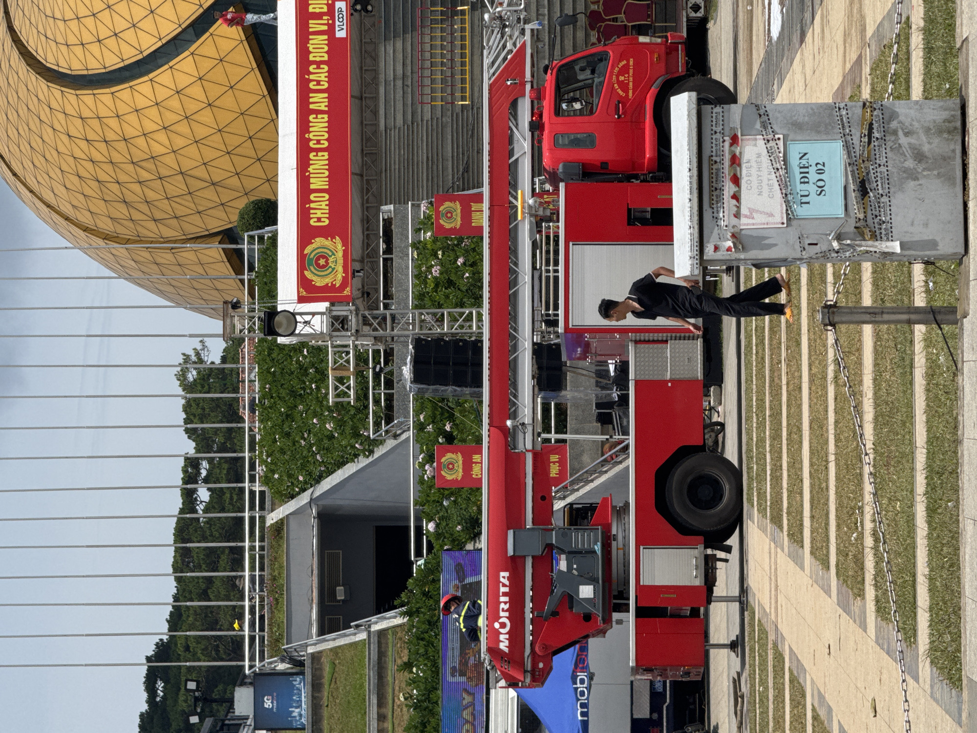 The image shows a red fire truck with a ladder extended, parked on a paved area with grass strips in front of a large, distinctive gold-domed building. The building appears to be the Da Lat Cultural Palace in Da Lat, Vietnam, given its architecture. A man in a black shirt and pants is walking away from the fire truck. In the background, there are stairs leading up to the building, and several people are visible on scaffolding or elevated platforms near the structure. A large red banner with Vietnamese text and official emblems is displayed above the fire truck. Another smaller banner with the word "CÔNG AN" (Police) is also visible. There is a gray electrical box in the foreground, taped with warning labels. The environment suggests an outdoor event or preparation for one.