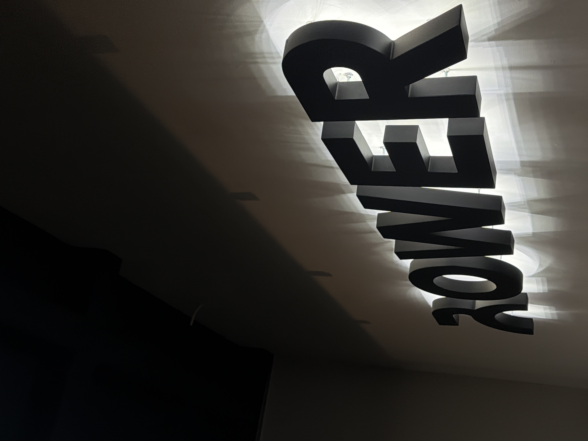 The photograph captures a vertical sign that spells out the word "POWER," each letter constructed in a three-dimensional, block-like design and internally illuminated to create a halo effect against the surface it is mounted to. The letters are large and prominent, casting shadows and reflecting light onto the surrounding ceiling and walls. The sign is likely in Puebla, Mexico, as the prompt asks. The overall ambiance suggests an indoor setting, possibly an office, studio, or retail space where branding and visual appeal are important. The left side of the frame is darker and appears to show the corner of a wall or piece of furniture, contrasting with the brightly lit sign. The entire image is shot from a low angle, emphasizing the size and presence of the sign.