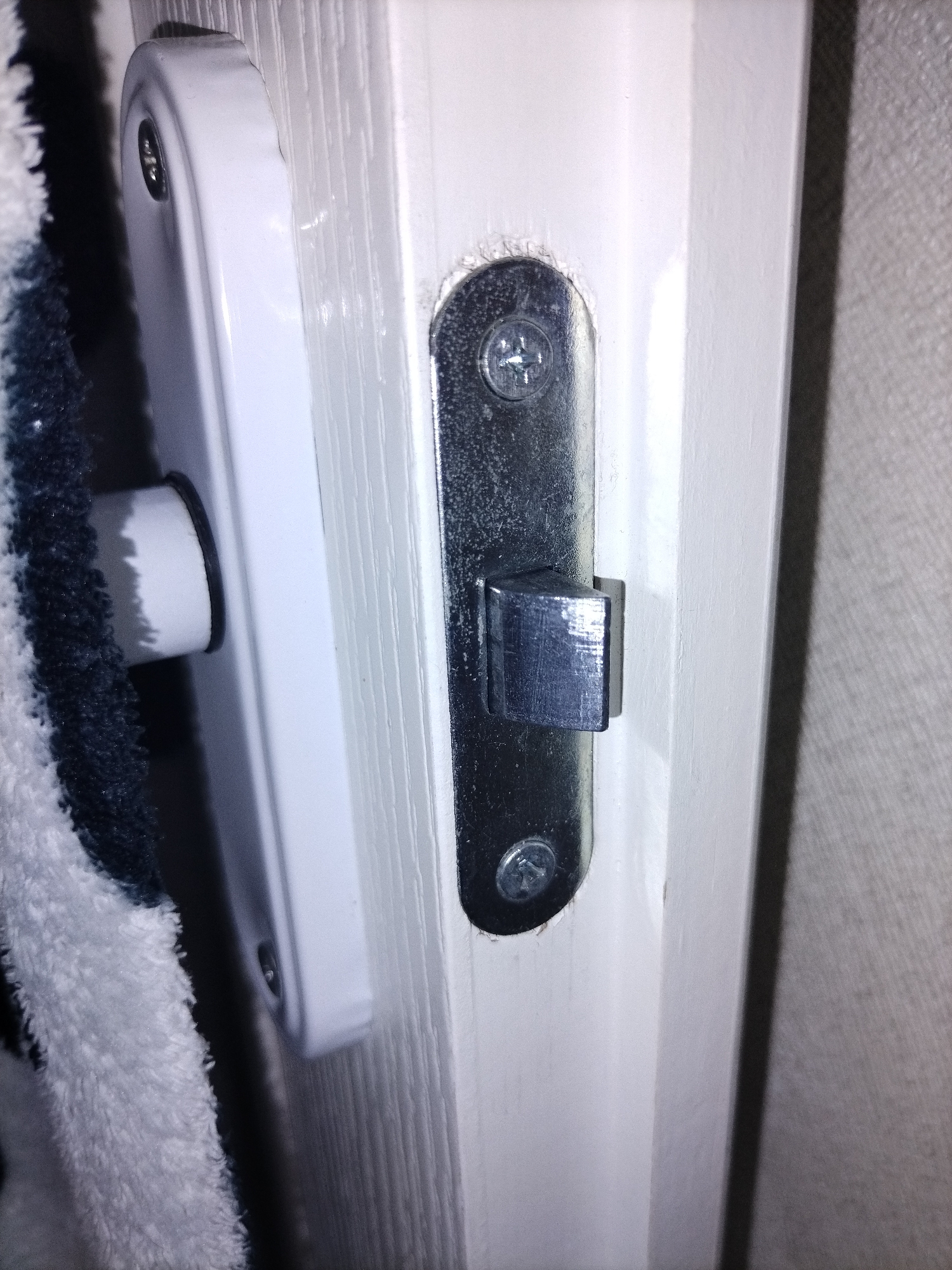 A close-up, eye-level, slightly angled shot shows a white door frame with a white strike plate and a metal latch mechanism. A dark blue and white striped towel is partially visible on the left side of the frame, obscuring part of the door and strike plate. The strike plate is attached to the door frame with two visible silver screws. The metal latch mechanism is embedded within the door frame and features a silver-colored tongue that is extended. The background on the right is a textured light gray wall. The lighting is dim, casting shadows on the scene.