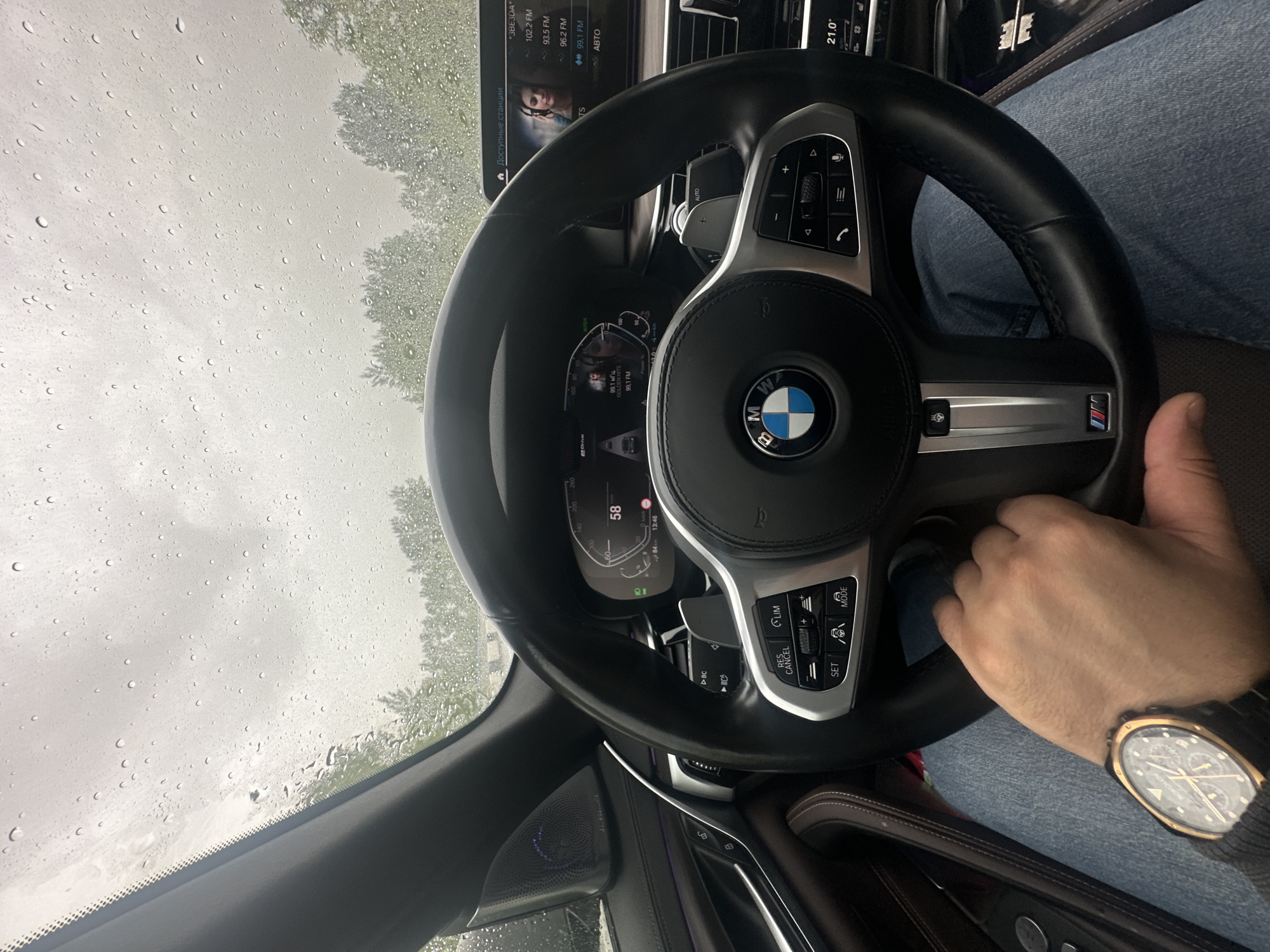 The photograph depicts an interior view of a BMW automobile, specifically focusing on the driver's perspective. The steering wheel is prominently centered, displaying the BMW logo and M badge. The driver's hand is visible holding the steering wheel, with a wristwatch on their wrist. The car's dashboard and infotainment screen are also partially visible. Through the windshield, raindrops are evident, suggesting wet weather conditions. The surrounding environment is blurred, hinting at a tree-lined area or urban setting. The car's speedometer shows a reading of 58, and the infotainment system displays radio frequencies and station names. The interior of the car features leather or similar premium materials.