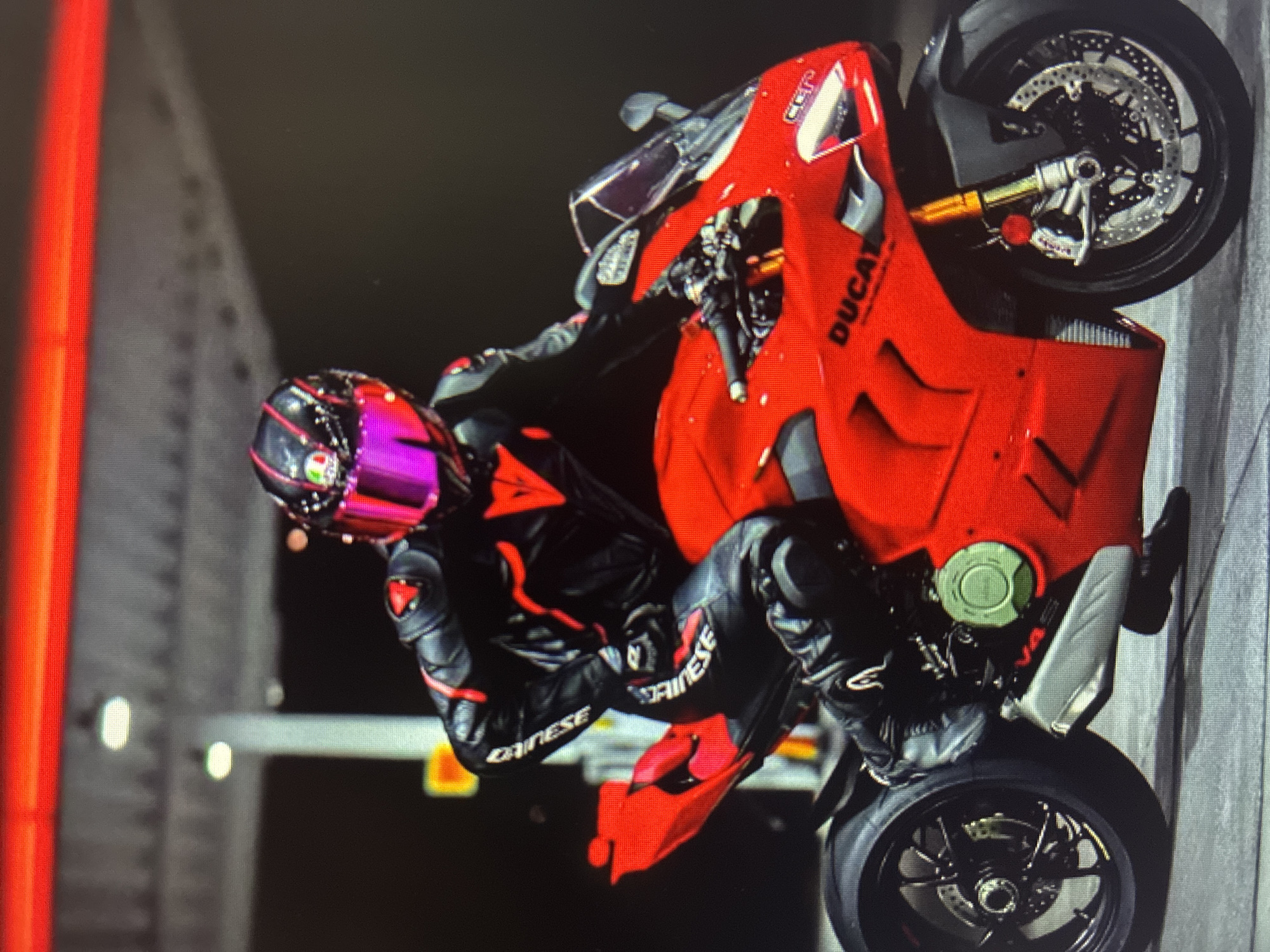 This dynamic, low-light image, possibly taken in Asenovgrad, Bulgaria, captures a person performing a wheelie on a bright red Ducati motorcycle. The motorcycle is angled sharply upwards on its rear wheel, with its front wheel lifted high off the ground. The word "DUCATI" is clearly visible on the side fairing of the bike, which also features black wheels, exposed disc brakes, and a notable green circular component on the engine block.
The rider is fully clad in a black leather racing suit with distinctive red accents, bearing the brand name "DAINESE" on the arms and legs, signifying high-performance gear. Their head is protected by a vibrant, iridescent or metallic purple and pink full-face helmet. The rider's body language suggests active engagement and skill in controlling the powerful machine during the stunt.
The background is predominantly dark, indicating night-time or very low-light conditions. On the left side of the frame, a prominent vertical red light source extends upwards. Further in the background, a large, grey, structured element with vertical lines and horizontal beams is visible, hinting at an industrial setting, a bridge, or a similar large construction. The image conveys a sense of speed, skill, and daring.