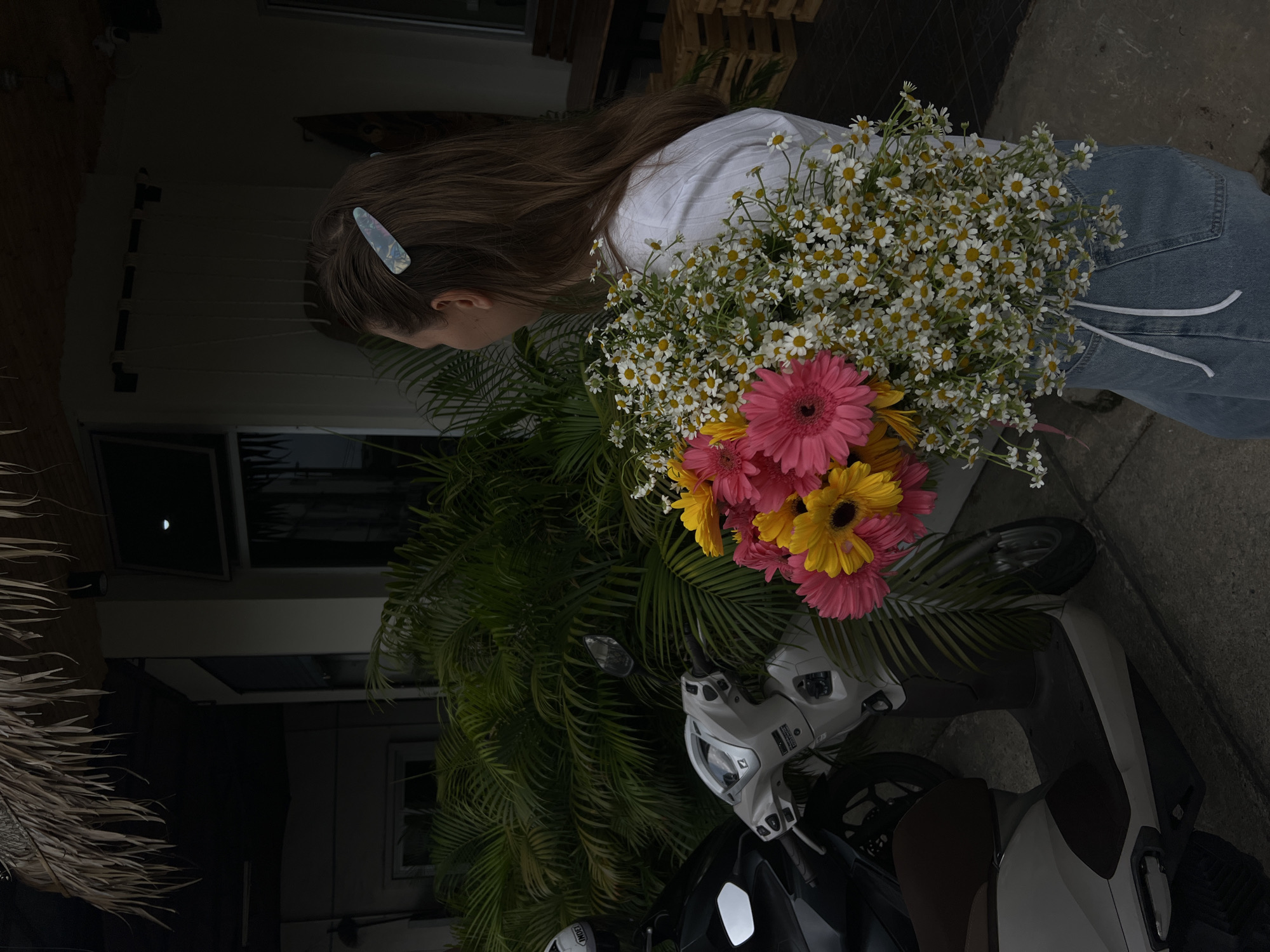 In Phuket City Municipality, Thailand, an image shows a woman holding a large bouquet of flowers. The bouquet consists of daisies and pink and yellow gerbera daisies. The woman has long brown hair and a blue hair clip. She wears a white shirt and blue jeans. A white scooter is visible in the background, as well as palm trees and a building.