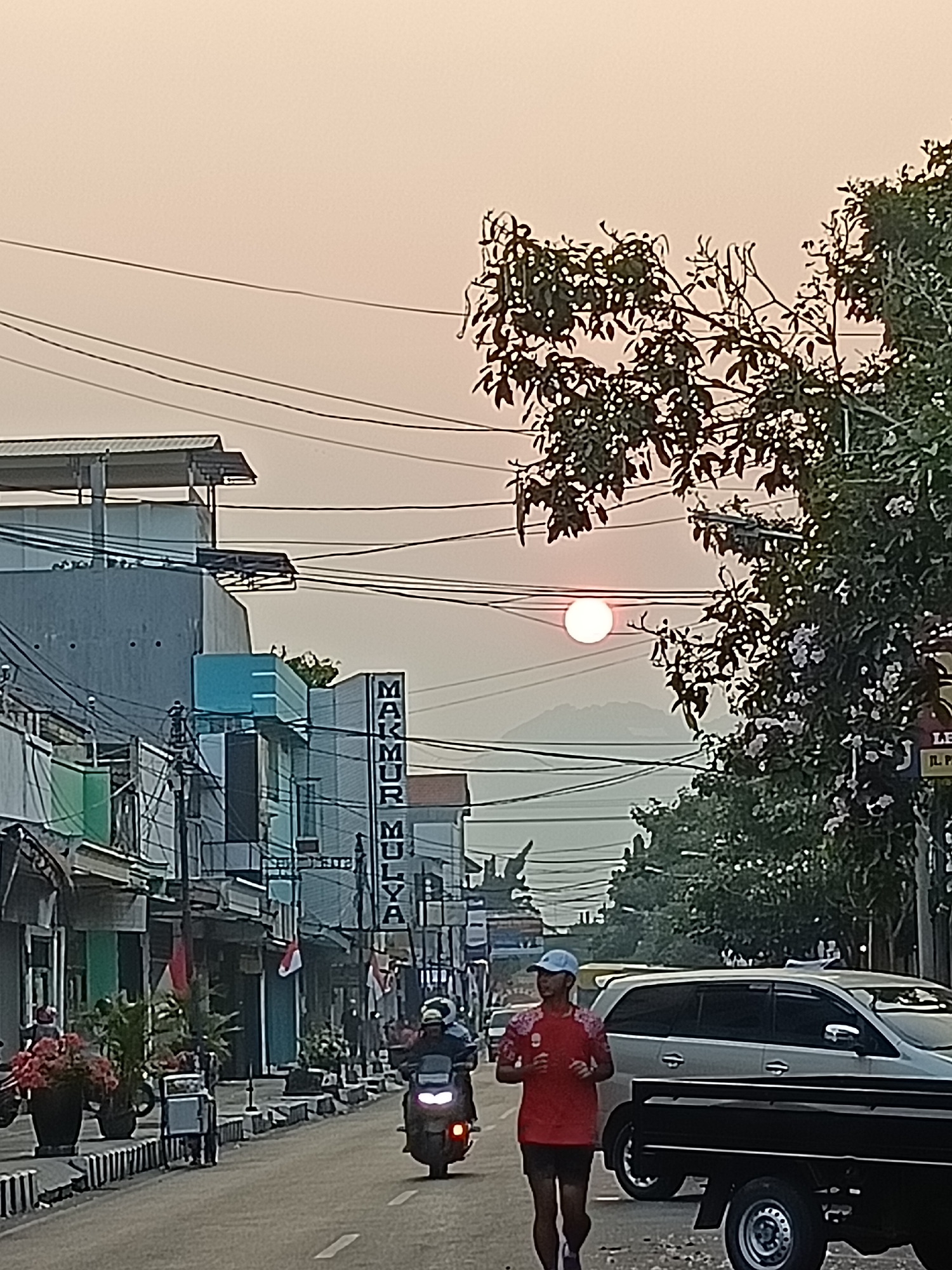 A street scene in Kediri City, Indonesia, features a person jogging on the right side of the road. The jogger, wearing a red shirt and black shorts with a light blue cap, is mid-stride. Behind the jogger, a silver SUV is parked on the left side of the road, and a black truck is partially visible in the foreground. The road is lined with buildings and shops on the left, some with awnings and signage, including one that reads "MAKMUR MULYA." There are also several motorcycles on the road, one of which is moving towards the viewer on the right. Power lines crisscross the sky, and trees with dark leaves and some white flowers are visible on the right side of the image. The sun is visible in the hazy sky, casting a soft glow over the scene, and mountains are faintly visible in the distance. The image appears to be taken during sunrise or sunset due to the warm light and hazy atmosphere.