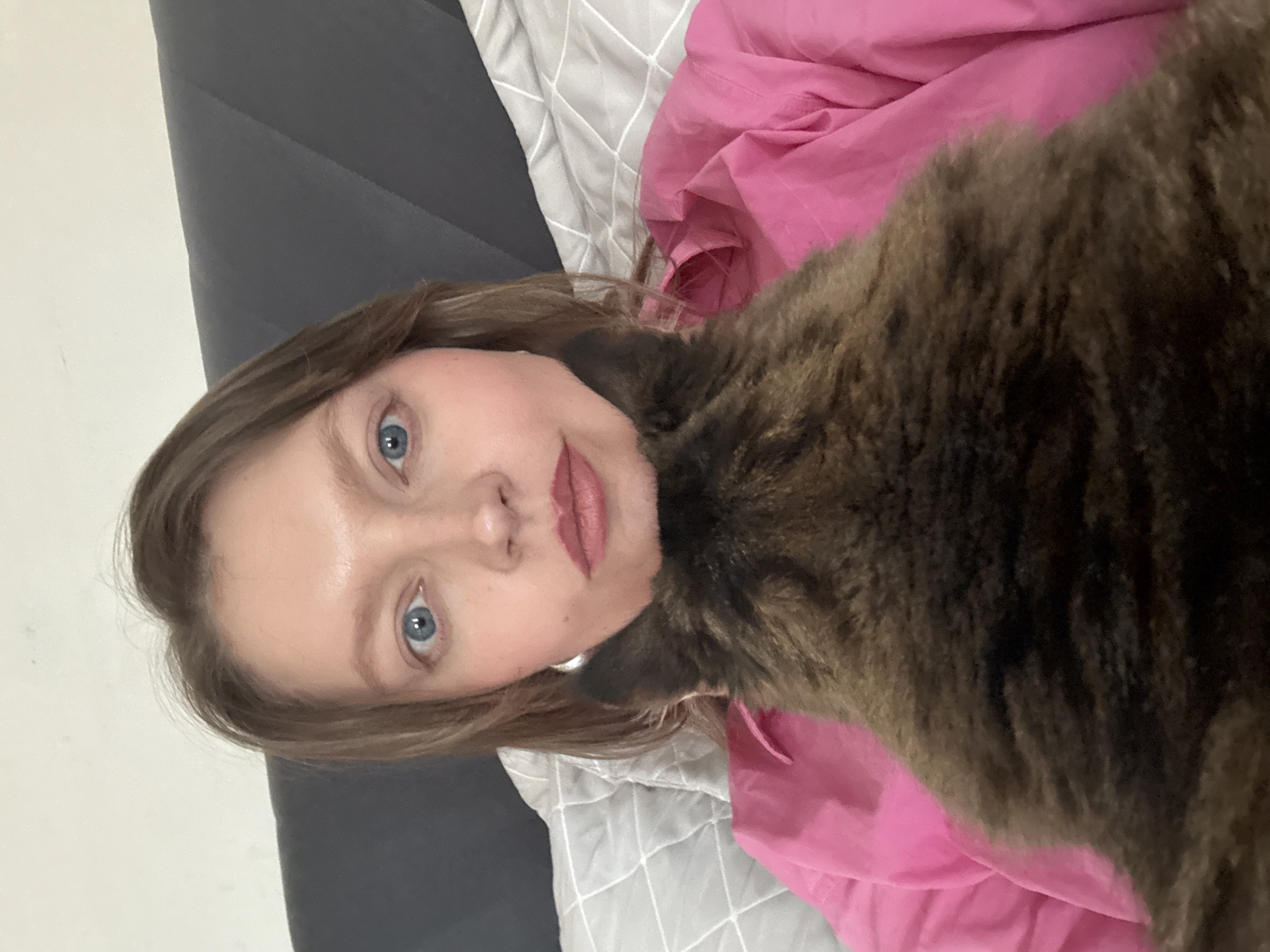 A woman with light skin, blue eyes, and light brown hair lies on a bed. She is partially covered by a pink sheet and a gray comforter with a white grid pattern. A large, brown tabby cat rests on or next to her, partially obscuring her face. The setting appears to be indoors, likely a bedroom, and the lighting suggests daytime. There is no indication of the location being Izhevsk, Russia.