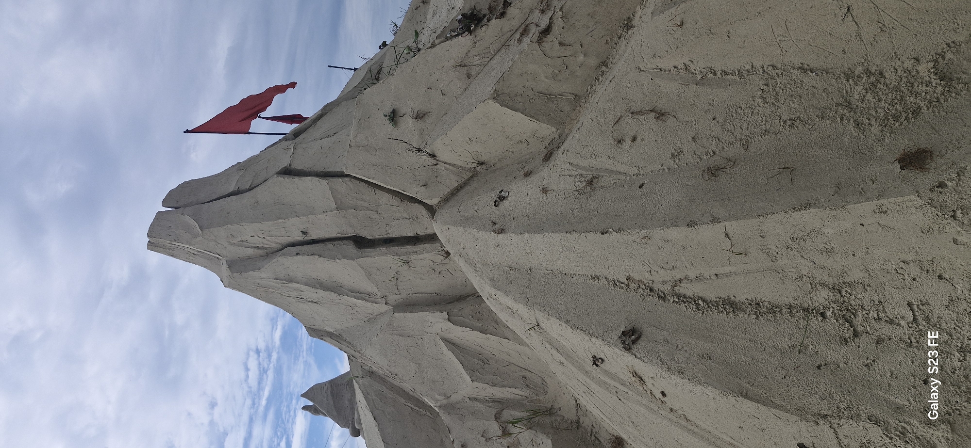 The image displays a large sand sculpture resembling a mountain with jagged peaks. Two red flags are planted at the summit, with one larger flag prominently displayed. The sculpture is detailed, with visible cracks and lines suggesting geological formations. Sparse vegetation, such as dry grass and leaves, is present on the sandy slopes. In the background, a smaller sand structure is visible to the left, and a hazy sky with scattered clouds forms the backdrop. The lower left corner of the image contains a watermark that reads "Galaxy S23 FE". The image does not depict the city of Jelgava, Latvia.