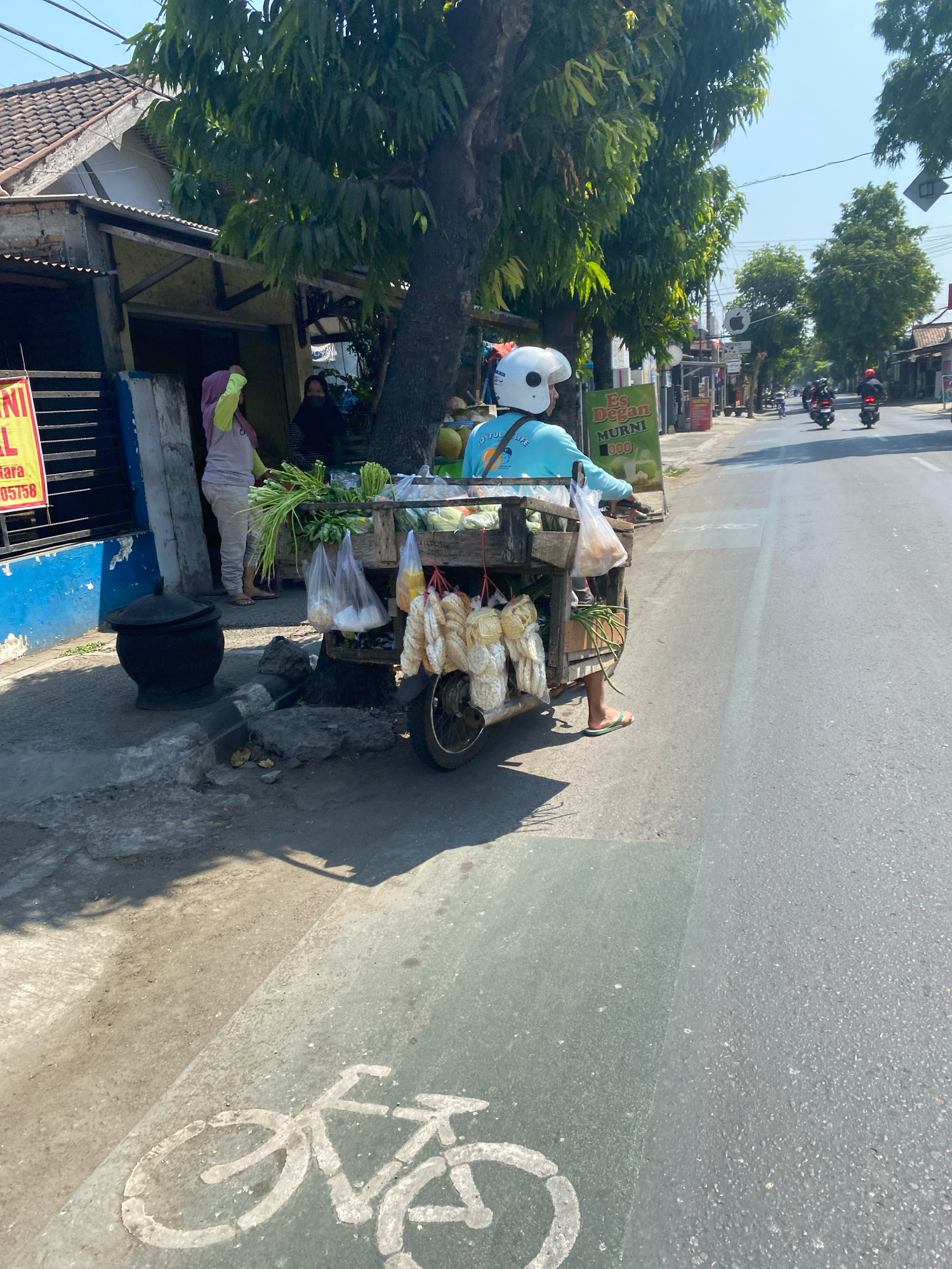 In Kediri City, Indonesia, a street vendor on a motorcycle is selling vegetables and snacks. The vendor, wearing a blue shirt and white helmet, is positioned on the right side of the road, near a row of buildings and trees. The motorcycle is equipped with a wooden cart filled with produce. Two women are standing near a stall on the left side of the road, one of whom is wearing a pink headscarf and holding a green bag. The road surface features a painted bicycle symbol, indicating a bike lane. The weather appears to be sunny with a clear sky.