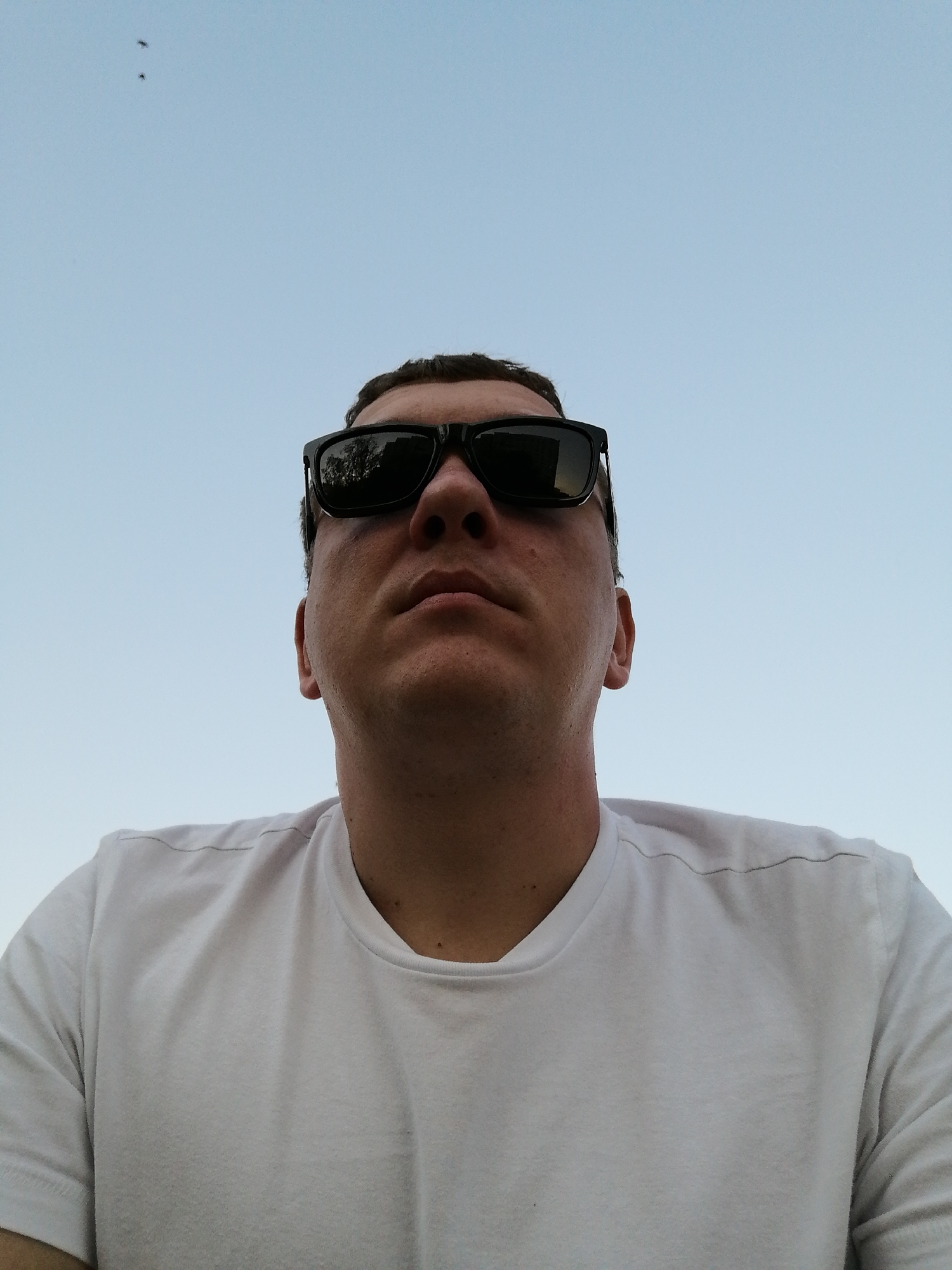A low-angle, eye-level photograph features a person wearing a white t-shirt and black sunglasses. The subject is looking up. The background is a light blue sky. Two small, dark objects are visible in the upper left corner of the frame. The location is Unknown, Unknown.