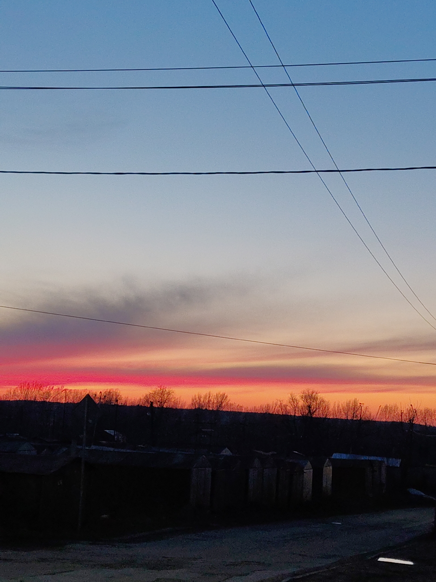 The image shows a twilight scene in an urban area. The sky transitions from blue at the top to pink and orange near the horizon. Power lines are visible across the top of the frame. Below the horizon, there's a dark silhouette of trees and rooftops. In the foreground, there are several dark, rectangular structures, possibly garages or sheds, lining a road or open area. No people are visible. The location is Unknown, Unknown.