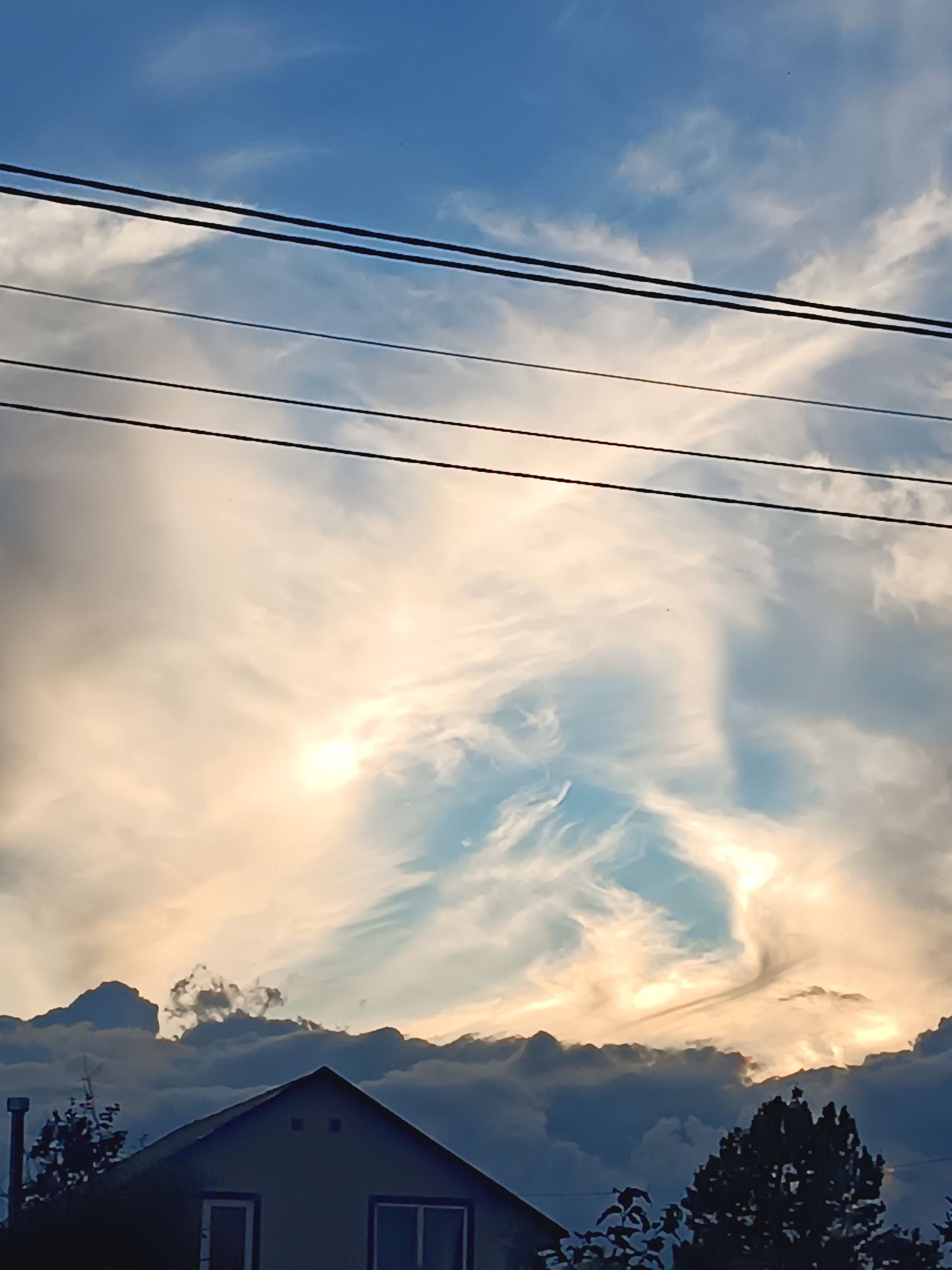 This is a photograph taken in Novaya Derevnya, Russia, showing a house with a dark-colored roof in the foreground. Above the house, a dramatic sky is visible with a mix of dark and light clouds. The sun appears to be partially obscured by clouds, casting a warm, golden light on the sky. Multiple power lines stretch horizontally across the upper portion of the image. Trees are also visible on the left and right sides of the house.