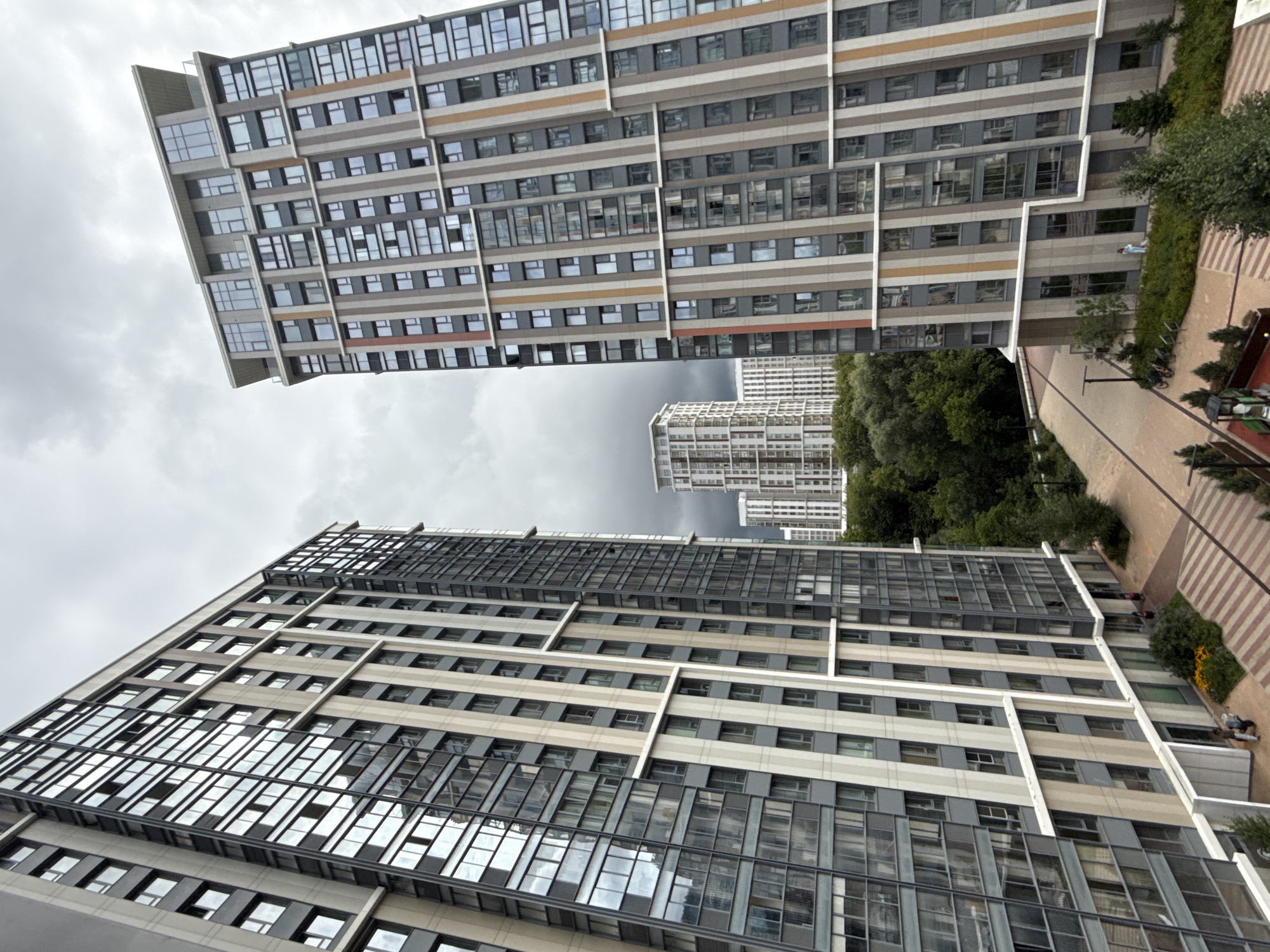 In Khimki, Russia, on a cloudy day, two large, modern residential buildings dominate the foreground. The building on the left is angled towards the viewer, its facade a mix of dark grey panels and large windows that reflect the overcast sky. The building on the right is also tall and rectangular, featuring vertical stripes of grey, beige, and thin orange and red accents, interspersed with numerous windows. Between these two structures, a middle ground features more residential buildings of similar modern architecture, set against a backdrop of dense green trees. In the foreground at the base of the buildings, paved pathways are visible, with some green landscaping and a few people walking. Bicycles are parked near one of the pathways. The overall scene depicts a contemporary urban residential environment.