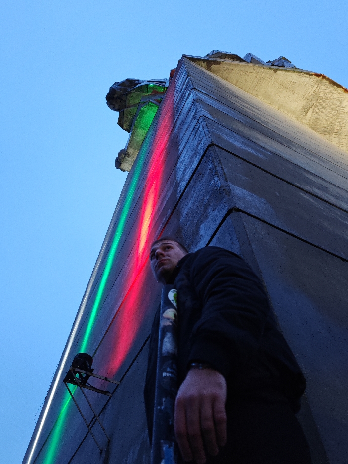 The image depicts a low-angle view of a brutalist concrete monument, identified as the "Monument to 1300 Years of Bulgaria" in Shumen, Bulgaria. A male subject stands in the lower right quadrant, positioned next to a dark pole with visible markings. He is looking upwards, towards the monument's illuminated facade.
The monument's surfaces are grey, textured concrete, with sharp angles and layered sections. A stylized lion's head, part of the monument's sculpture, is partially visible at the upper left edge, illuminated with a greenish light. Two prominent vertical strips of artificial light, one green and one red, run along a vertical edge of the monument, indicating an LED lighting installation. A small, black spotlight is mounted on a metal bracket near the bottom left, directed upwards.
The male subject, a young adult, has short dark hair and is wearing a dark jacket or sweatshirt. His right arm and hand are visible, with a dark bracelet on his wrist. The background consists of a clear, light blue sky.