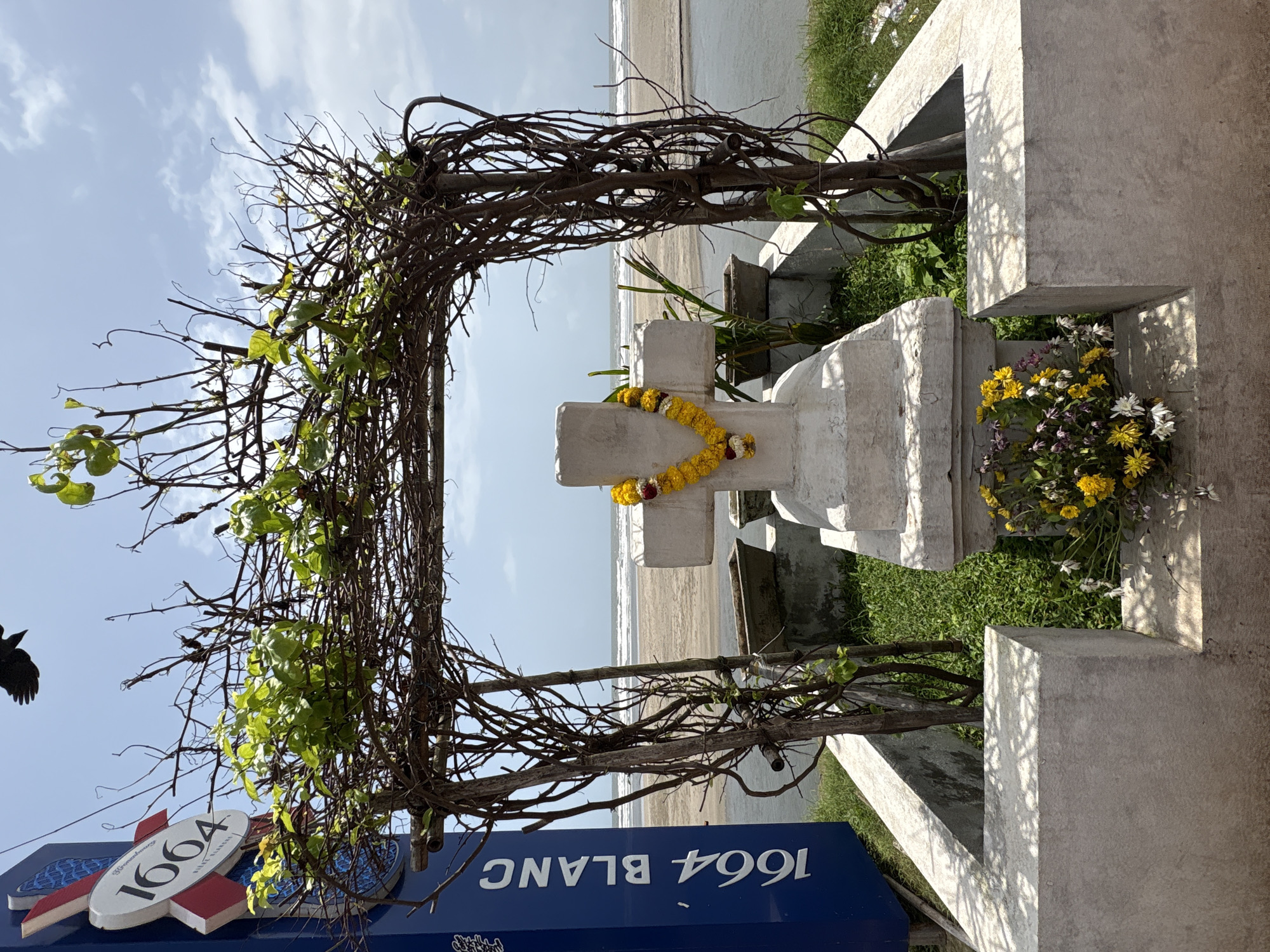 A white concrete cross, adorned with a marigold garland, stands in a small, enclosed garden on a beach. The cross is the central focus, framed by a rustic archway made of intertwined branches. To the left of the cross, a blue sign with "1664 BLANC" in white lettering is visible, suggesting a nearby establishment or landmark. In the background, the ocean meets the sandy shore under a partly cloudy sky. Small potted plants are placed within the enclosure around the cross. A bouquet of yellow and white flowers rests at the base of the cross. The overall scene depicts a memorial or decorative element situated in a coastal environment.