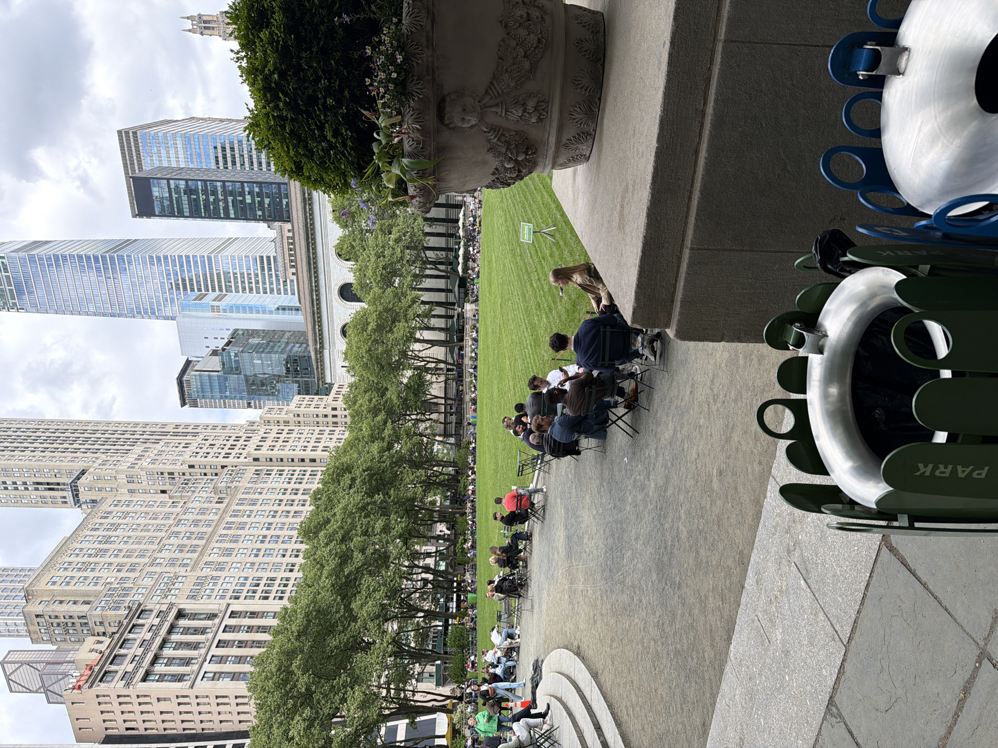 The image depicts Bryant Park in New York City, United States. A large, green lawn occupies the center of the frame, with numerous people seated in rows of chairs. Trees line the edge of the lawn, providing shade. Several tall buildings surround the park, including modern skyscrapers and older, ornate buildings. The sky is overcast. A large stone planter with vegetation sits in the foreground, partially obscuring the view. In the lower right corner, a distinctive trash receptacle with the word "PARK" is visible. People are sitting, presumably watching something.