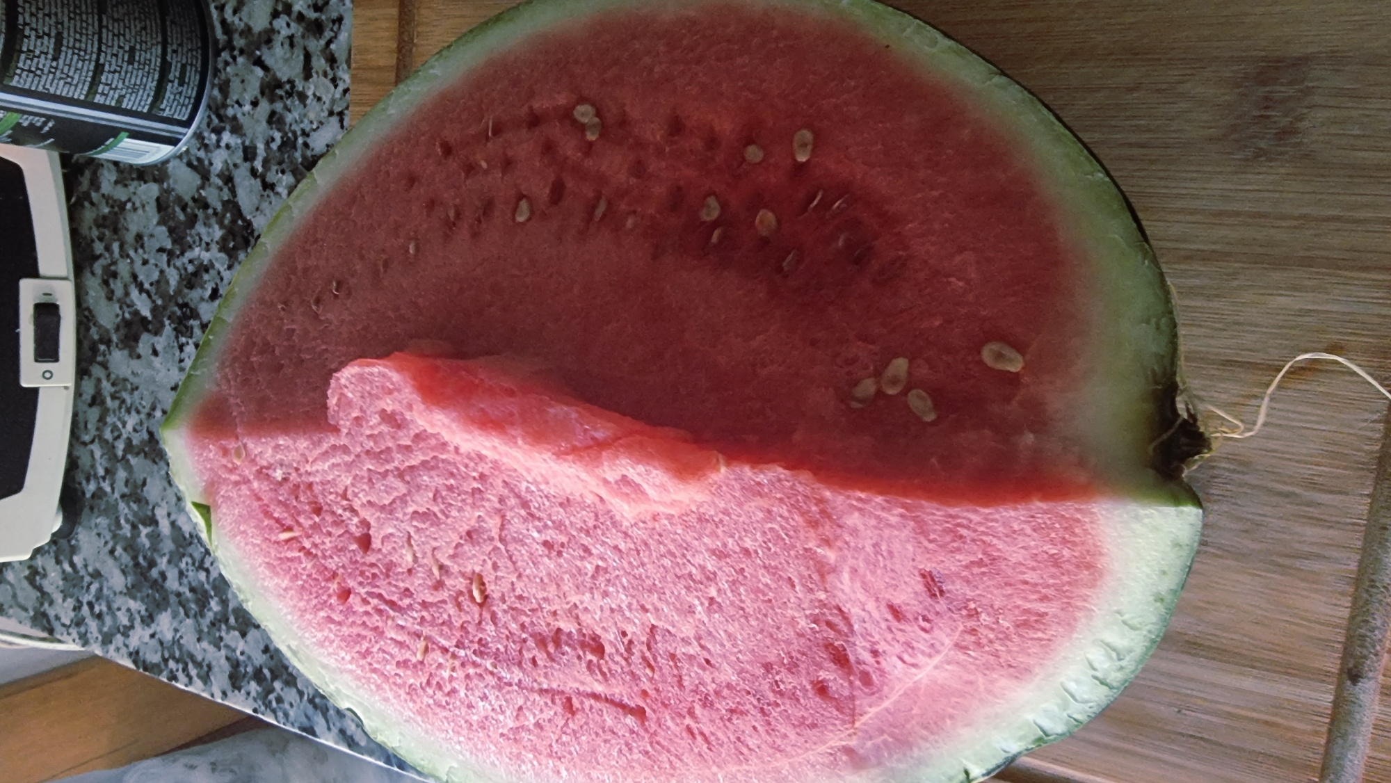 A large slice of watermelon is displayed on a wooden cutting board. The watermelon has a vibrant pink flesh and a green rind. A smaller piece of watermelon has been cut and placed on top of the larger slice, creating a layered effect. The surface of the watermelon is textured with small pores and scattered black seeds. The cutting board shows visible wood grain. In the background, a speckled countertop and a white appliance are partially visible. The scene is well-lit, with the light source appearing to be from the top left, casting soft shadows. The image is a close-up shot, focusing primarily on the watermelon. The location in the city of Sant Joan Les Fonts, Spain is not depicted or inferable from the image itself.