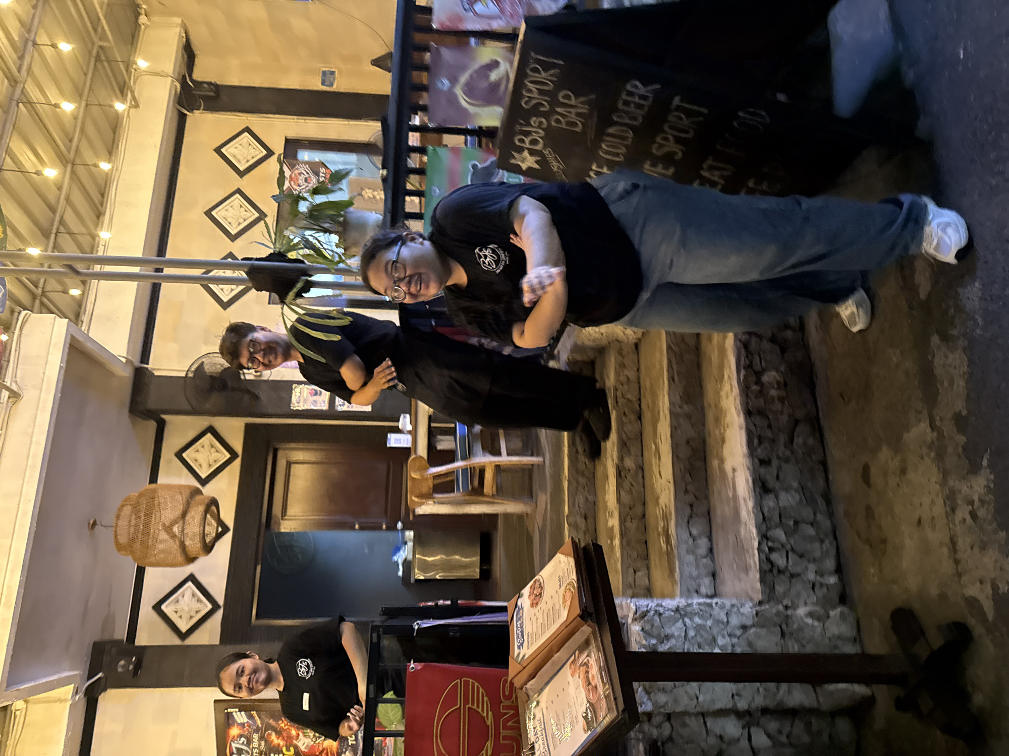 In Denpasar, Indonesia, at an establishment identified as "BJ's Sport Bar," three individuals are present. Two women and one man, all appearing to be staff members due to their uniform black t-shirts, stand on a stone staircase. The woman on the left, with dark hair tied back, is positioned by a red sign with a circular logo. The man in the center, wearing glasses and a black t-shirt, stands higher on the stairs, facing the viewer. The woman on the right, also wearing glasses and a t-shirt with the "BJ's" logo, is positioned at the bottom of the stairs, facing the viewer with her arms crossed. In front of her, a chalkboard sign displays "BJ's SPORT BAR," "COLD BEER," "VE SPORT," and "EAT FOOD." The environment suggests an outdoor or semi-outdoor seating area, with hanging lights and bamboo-style decor visible. Menus are displayed on a stand to the left of the stairs.