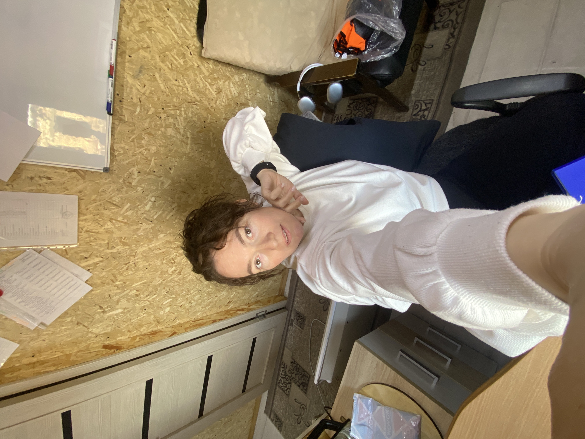 The image depicts an interior room, likely an office or workspace, located in Ol'khovskoye, Russia. A female individual with medium-length curly brown hair, dressed in a white long-sleeved top with voluminous sleeves and dark lower garments, is seated in a dark office chair. She extends her left arm towards the camera, holding a device for a selfie, and a black wristband is visible on her left wrist. The room's walls are finished with light-colored oriented strand board (OSB). In the upper left quadrant, a light-toned wooden door with two prominent dark horizontal stripes is visible, positioned above a patterned rug that covers the floor. Various documents are affixed to the OSB wall, adjacent to a white dry-erase board equipped with markers in its lower tray. A light-colored wooden desk, incorporating a drawer unit, occupies the lower left portion of the frame. To the right of the seated individual, silver over-ear headphones rest on a surface. Adjacent to the headphones, a clear plastic bag contains an orange object. A segment of beige upholstered furniture is visible in the right background. A blue light source is present at the bottom edge of the image.