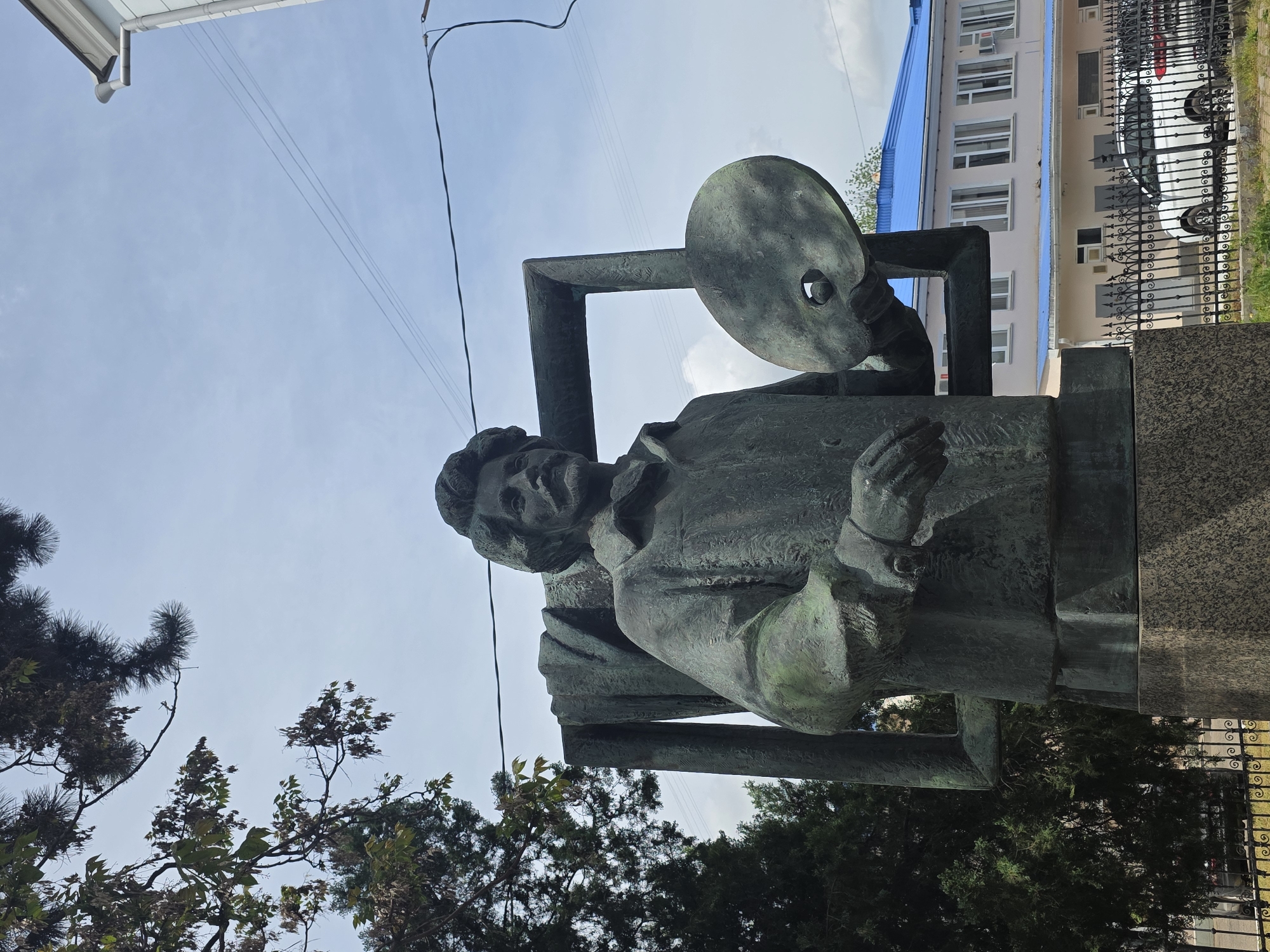 A statue of an artist, holding a palette, stands in Krasnodar, Russia. The bronze statue is set against a background of trees, a building with a blue roof, and a wrought iron fence. The sky is visible above.