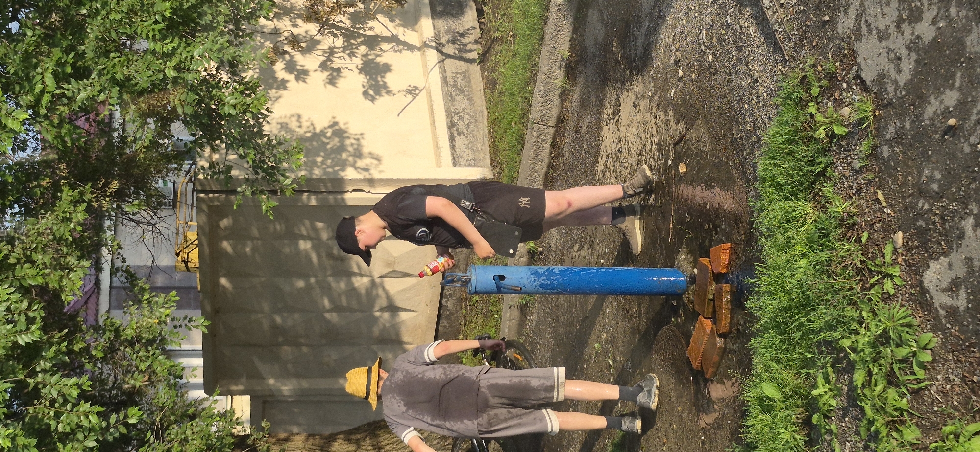 The image depicts two young males standing near a blue water pump on a sunny day. The male on the left, wearing a yellow hat, a gray t-shirt, and gray shorts, has his back to the camera. He appears to be wet, with visible water droplets on his t-shirt and shorts. The male on the right, wearing a black t-shirt, black shorts, and a black baseball cap, is holding a red and yellow bottle and looking at it. He is also wearing a black pouch attached to his shorts. They are standing on a wet, paved surface with some bricks arranged around the base of the water pump. Green grass and gravel are visible on the ground, and a tree with green leaves casts shadows on a light-colored wall in the background. The scene suggests an outdoor setting, possibly a park or a residential area, in Ussurijsk, Russia.
