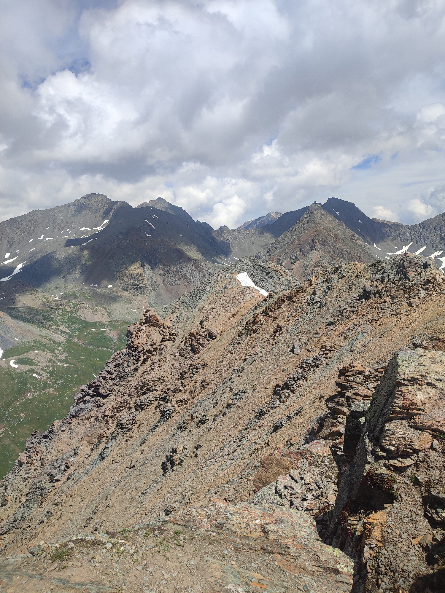 The image depicts a mountainous landscape under a cloudy sky. The foreground is dominated by a steep, rocky slope composed of loose scree and larger stones in shades of brown and grey. Several jagged rock formations are visible on this slope. In the middle ground, a similar rocky incline continues, with patches of snow scattered across it. Beyond this, further mountain peaks rise, some appearing sharper and more rugged, others smoother and more rounded. Patches of green vegetation are visible in the valleys between the mountains, interspersed with more rocky terrain and snow. The sky above is filled with a mixture of white and grey clouds, with glimpses of blue sky peeking through in some areas. The lighting suggests it is daytime, likely late morning or afternoon. The scene conveys a sense of rugged, natural wilderness. The provided location of Aktash, Russia, places this image within a region known for its mountainous terrain. There are no people, animals, or man-made objects visible in the image.