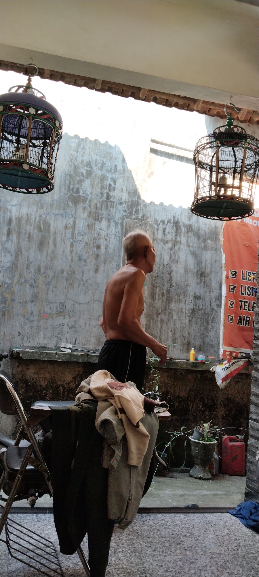 In Peganjaran, Indonesia, an elderly shirtless man with thinning white hair stands near a concrete wall, his back to the viewer. He wears black shorts. The scene appears to be an outdoor space, possibly a backyard or patio, with a textured, weathered concrete wall as a backdrop. Two decorative birdcages hang from the ceiling, one on the left and another on the right, each containing a bird. In the foreground, a metal folding chair is partially visible, with several items of clothing draped over it, including a beige sweater and dark pants. A small plant in a pot sits to the right of the man, near a red jerrycan. A poster with text, possibly advertising a business, is attached to the wall on the far right. The lighting suggests it is daytime, with sunlight casting shadows.