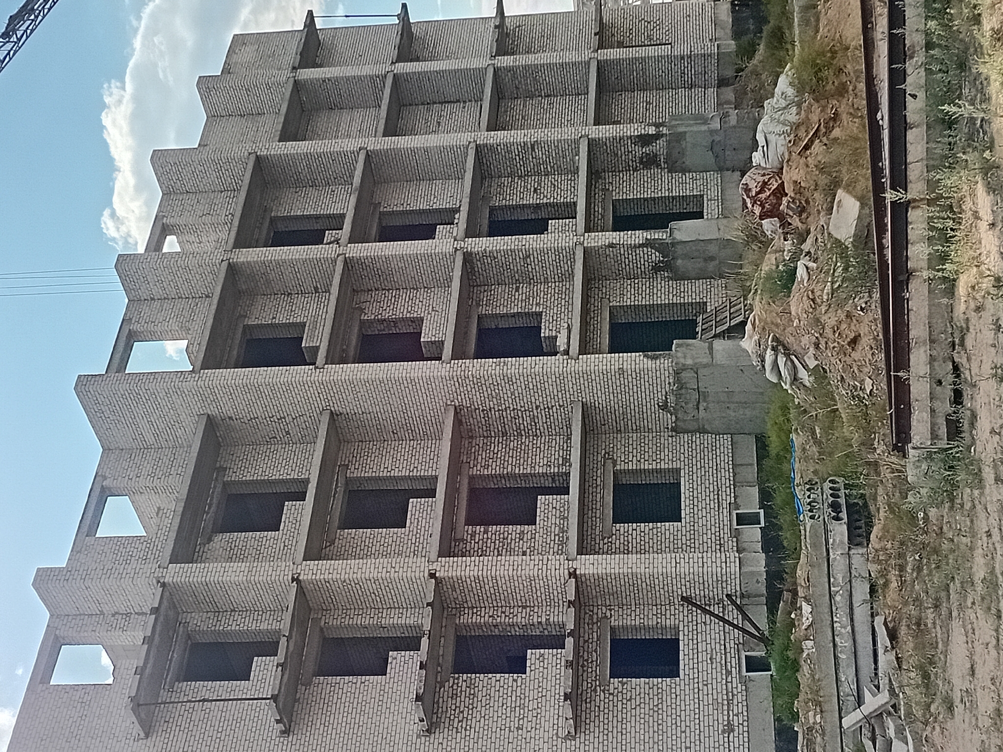 A partially constructed, multi-story brick building is the primary subject of this image, located in Semey, Kazakhstan. The building features multiple floors with open window and door frames, indicating it is in the early stages of construction. Concrete balconies or ledges jut out from each level. The exterior is constructed of light-colored bricks, and some areas show exposed concrete. In the foreground, stacks of concrete slabs and building materials are visible on the ground, along with piles of dirt and sand. Some rebar and metal structures are also present. The scene appears to be a construction site under a partly cloudy sky. There are no people visible in the frame.