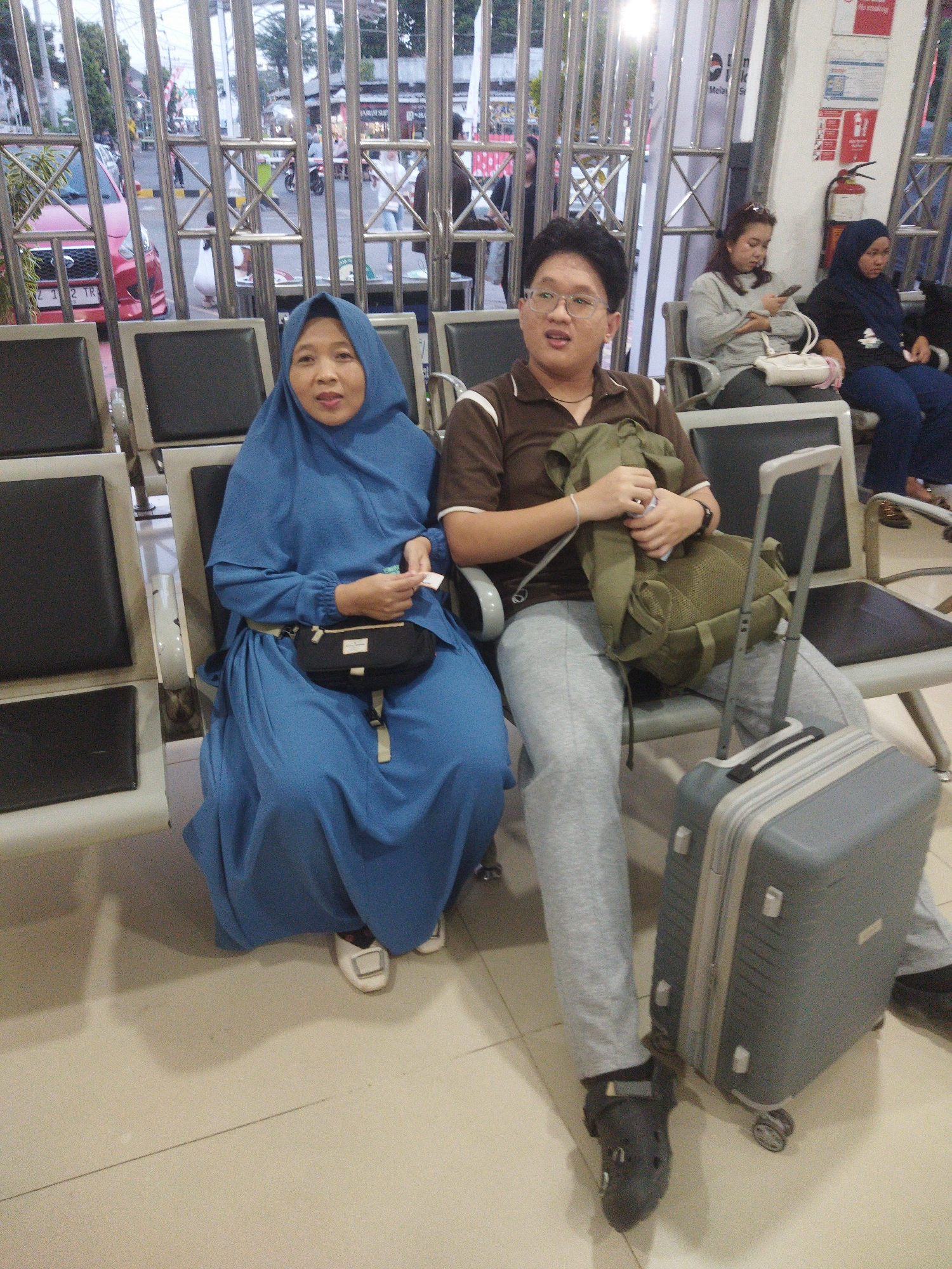 Two individuals are seated in what appears to be a waiting area, possibly a bus or train station, in the city of Banjar, Indonesia. The person on the left is a woman wearing a blue hijab and a long blue dress. She has a black pouch around her waist and is holding a small item in her right hand. The person on the right is a man wearing a brown polo shirt and grey sweatpants. He is wearing glasses and has a green backpack on his lap, which he is holding with his left hand. A grey rolling suitcase is positioned next to him. In the background, a red car is visible through a metal gate. Other individuals are seated further back in the waiting area.