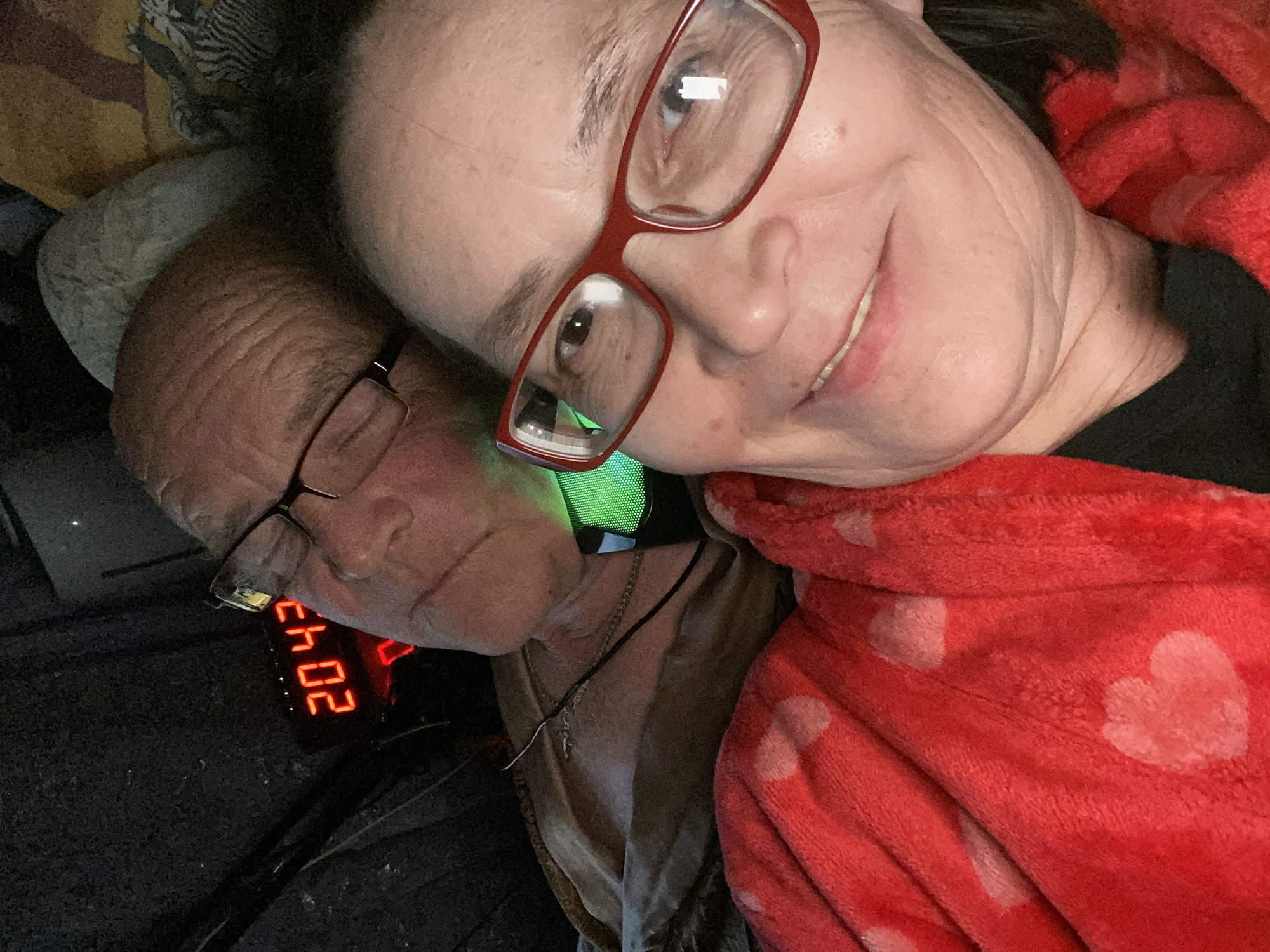 Two people are lying close together. One person, wearing red glasses, is turned toward the camera and smiling. The other person, wearing glasses, is facing away from the camera, and is close to a clock. The clock reads "20:43." A red blanket with white hearts is present. A necklace with a cross is visible. There is no indication in the image that it was taken in Novokuznetsk, Russia.