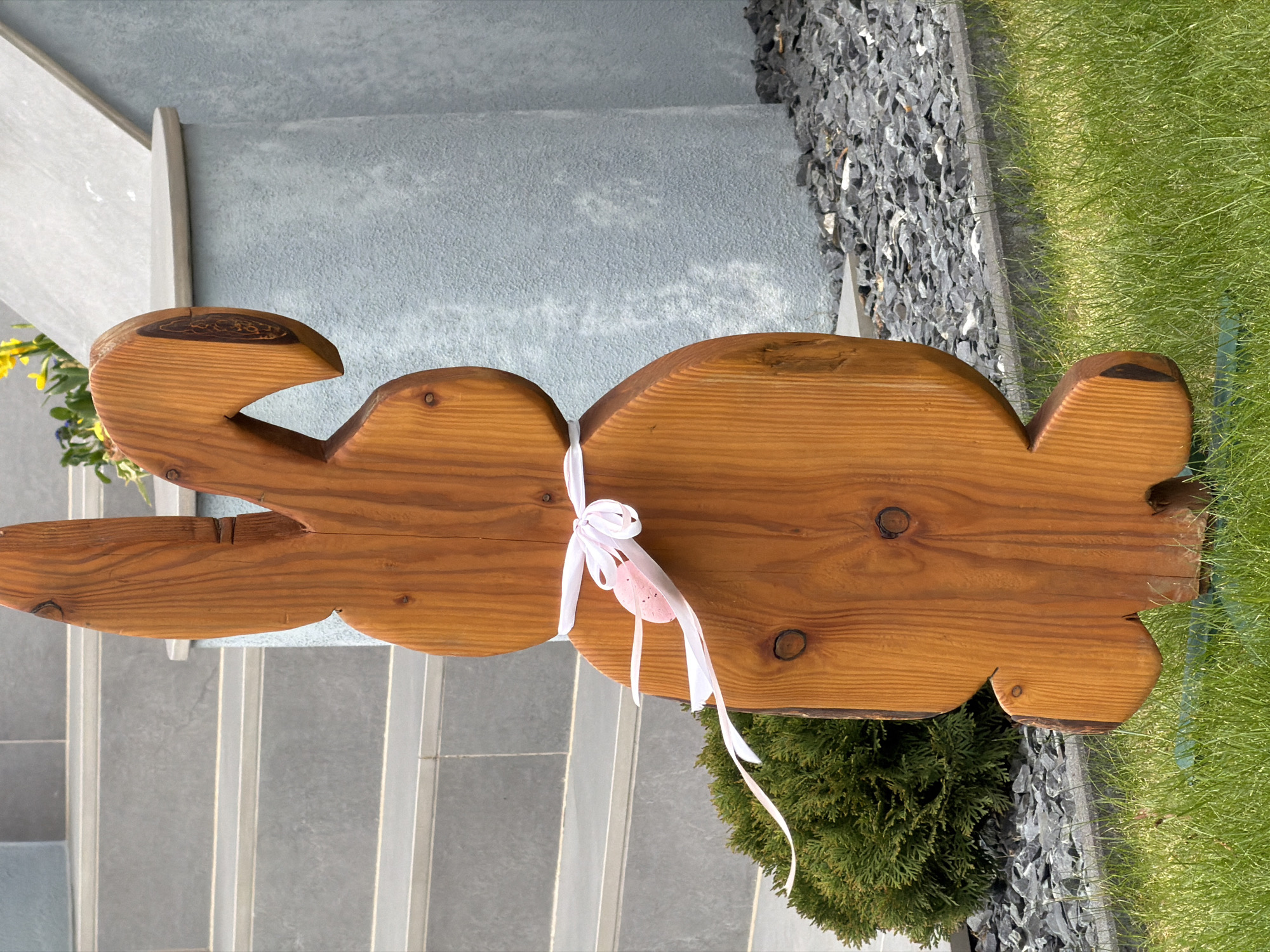 The image shows a wooden rabbit decoration tied with a white ribbon and a pink egg. The decoration is placed on grey stone steps. Next to the steps, there is a patch of green grass and a small evergreen shrub. The scene appears to be in an outdoor setting, possibly a garden or entrance area. The location is Esch-sur-Alzette, Luxembourg.