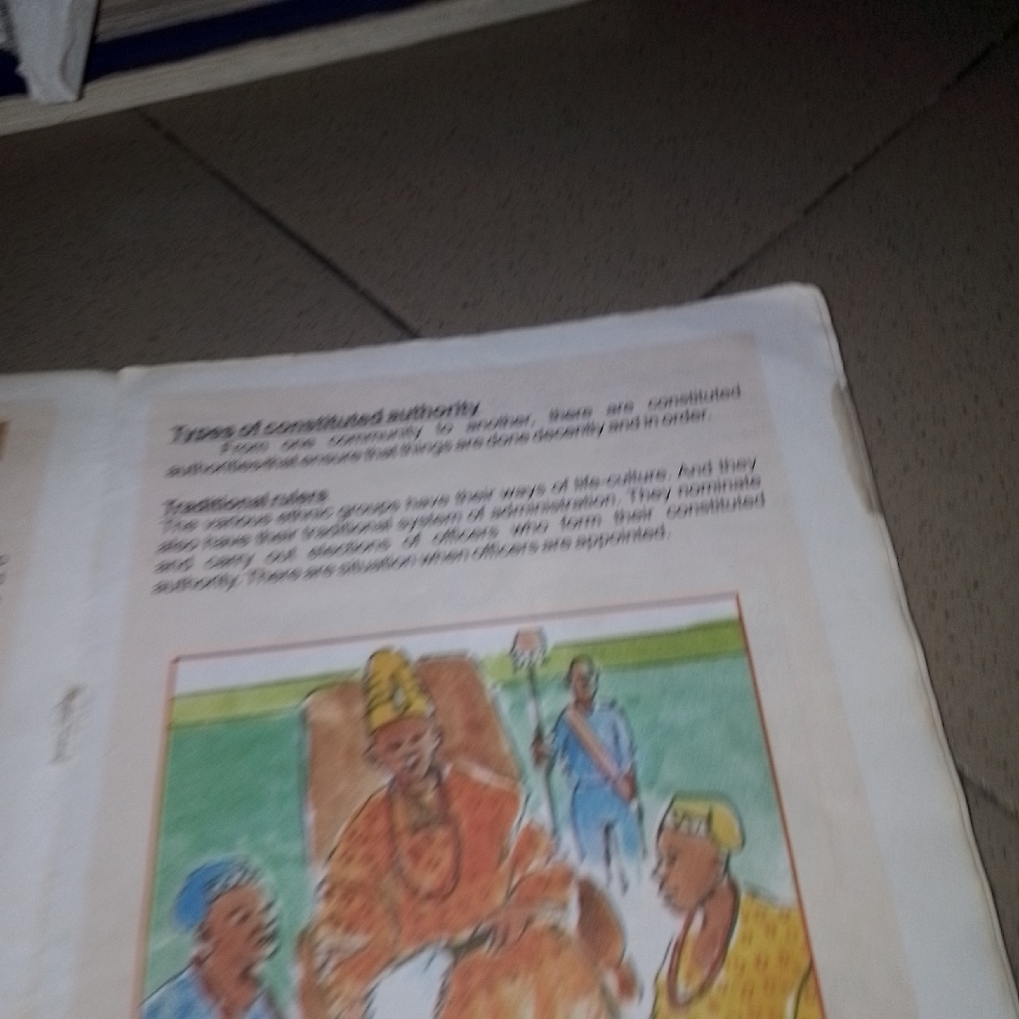 The image shows an open book on a textured, dark surface. The book displays text and an illustration. The text, in English, discusses "Types of constituted authority" and "Traditional rulers." The illustration depicts a scene with several figures. One central figure, wearing a yellow crown and an orange garment, appears to be seated. To the right and left of this central figure are other individuals, one wearing a blue garment and holding a staff, and two others wearing head coverings. The illustration is a watercolor-style drawing. The background of the book is white, with a green border around the illustration. The overall scene is a close-up of a page from a textbook or educational material.