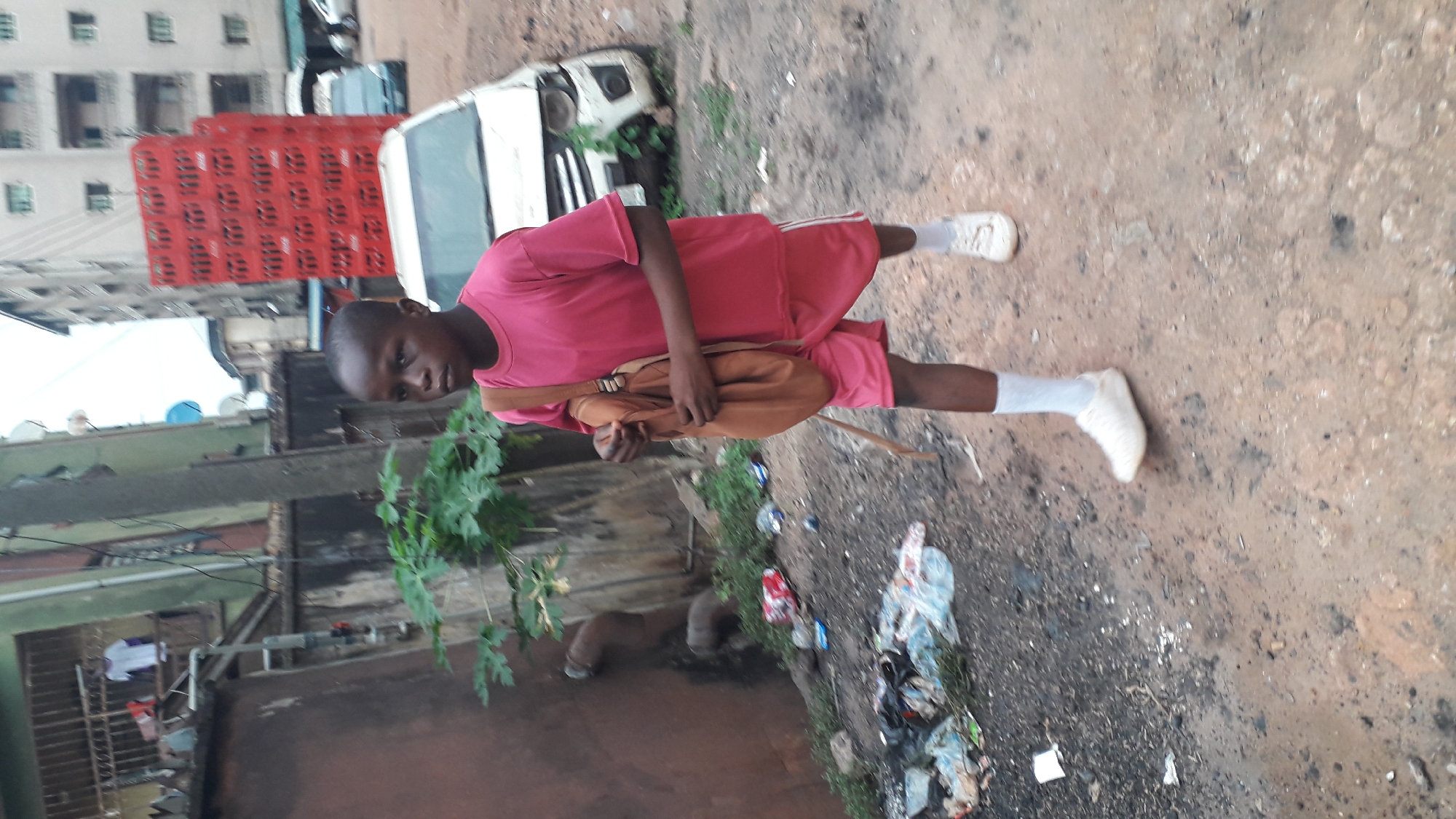 A young boy stands on a dirt and debris-strewn road in Obosi, Nigeria. He wears a red shirt and shorts, white socks and sneakers, and a brown bag is slung over his shoulder. In the background, a building is visible with red crates stacked in front of it, and a white vehicle is parked nearby. Rubbish is scattered on the ground near the boy. Vegetation grows along the road. A building with green framing and hanging laundry is also visible.