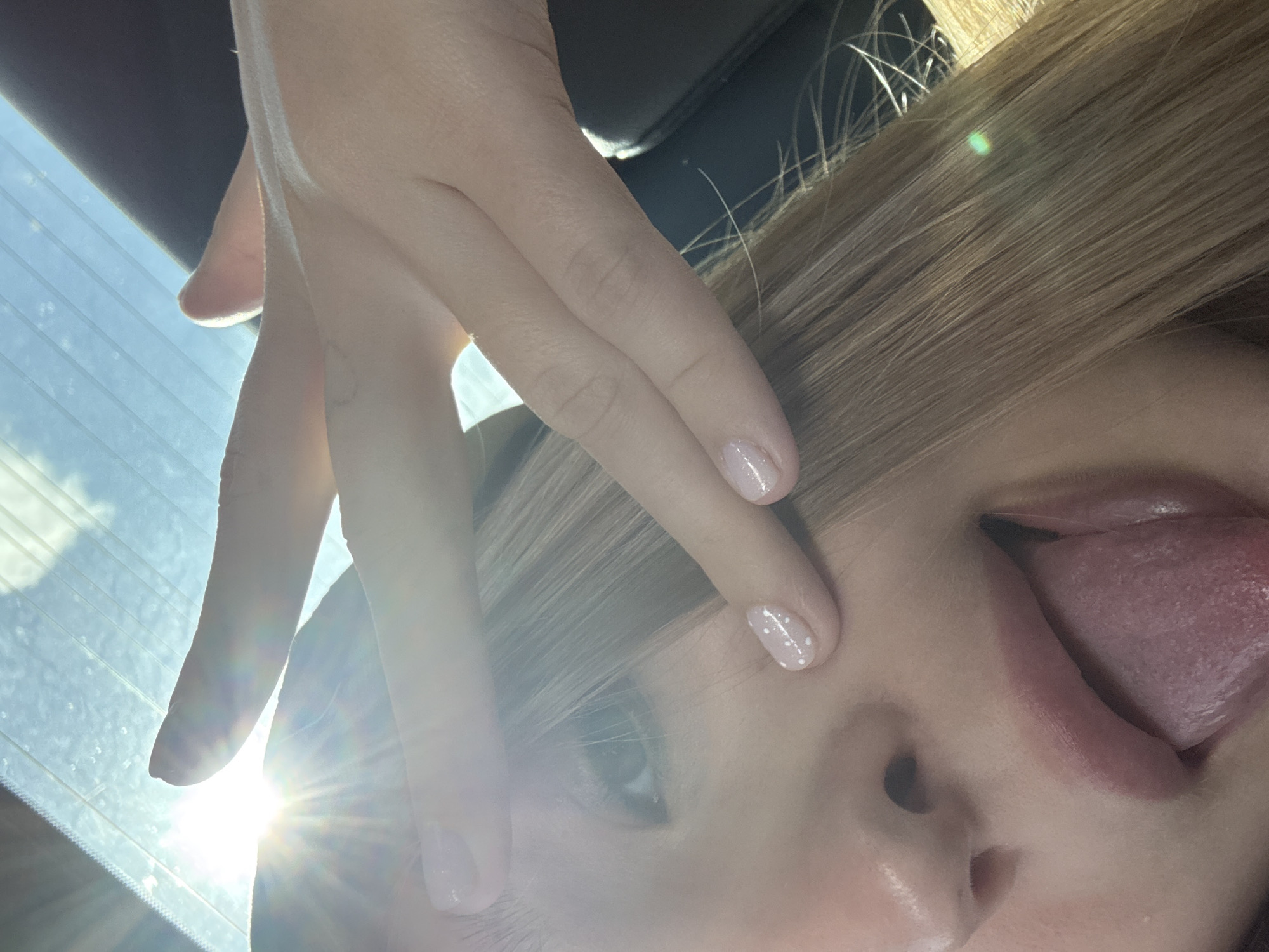 A close-up, low-angle shot shows a young woman's face, partially obscured by her hand and lens flare from the sun. She has light brown hair, and her tongue is sticking out. Her nails are painted a light pink color with small white dots. The background appears to be the inside of a car, with the sun shining through the windshield. There are no visible landmarks or indications of the city of Doksha, Russia.