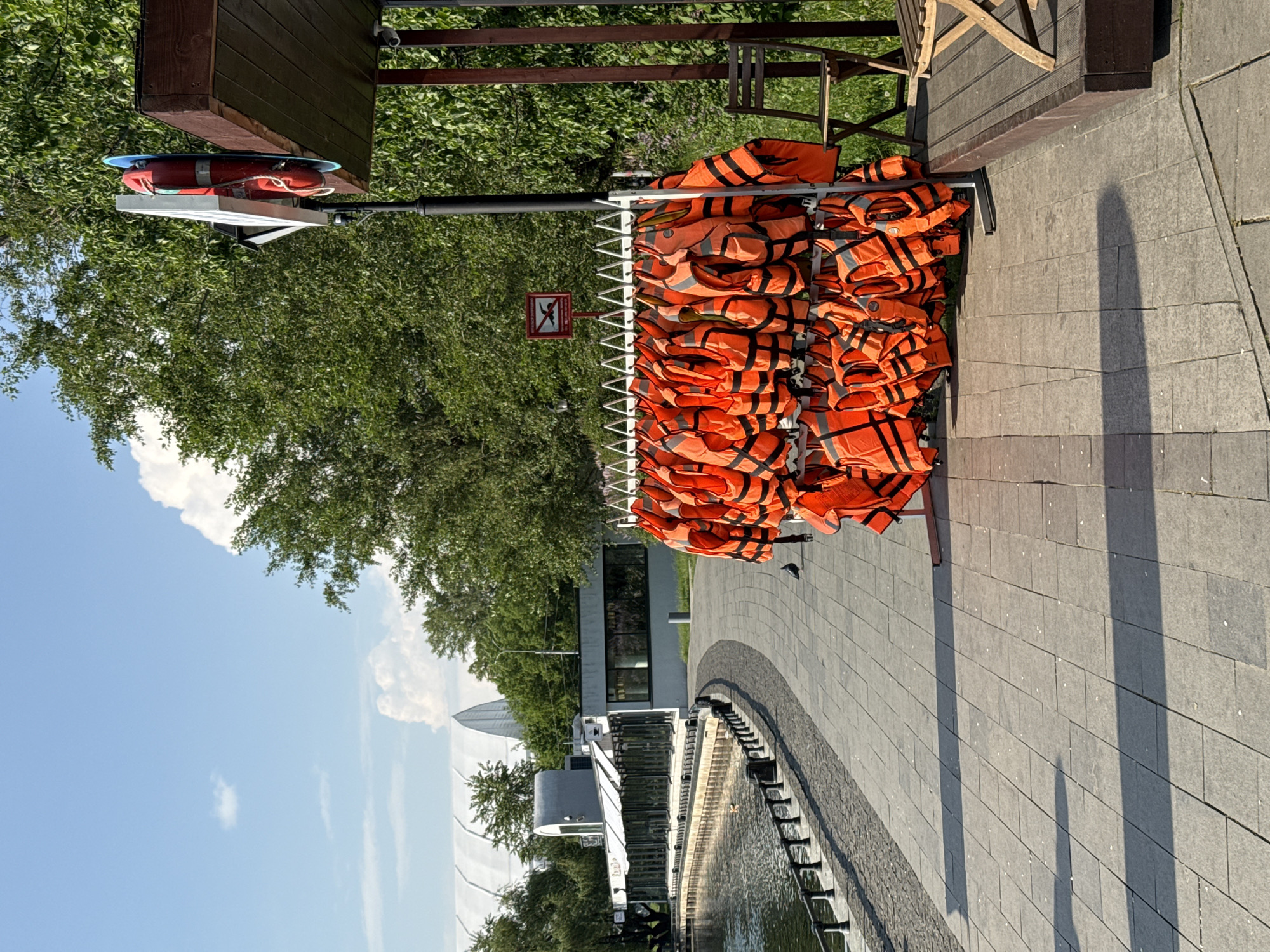 The image shows a rack of orange life vests, likely near a body of water. A life preserver is atop the rack. A small sign with an image of a person crossing out indicates it is a place to not jump into the water. The photo was taken near Gorky Park in Moscow, Russia. The sky is blue with white clouds, suggesting a sunny day. A brick walkway surrounds the water. In the background are trees and part of a building. The image is oriented in portrait mode.