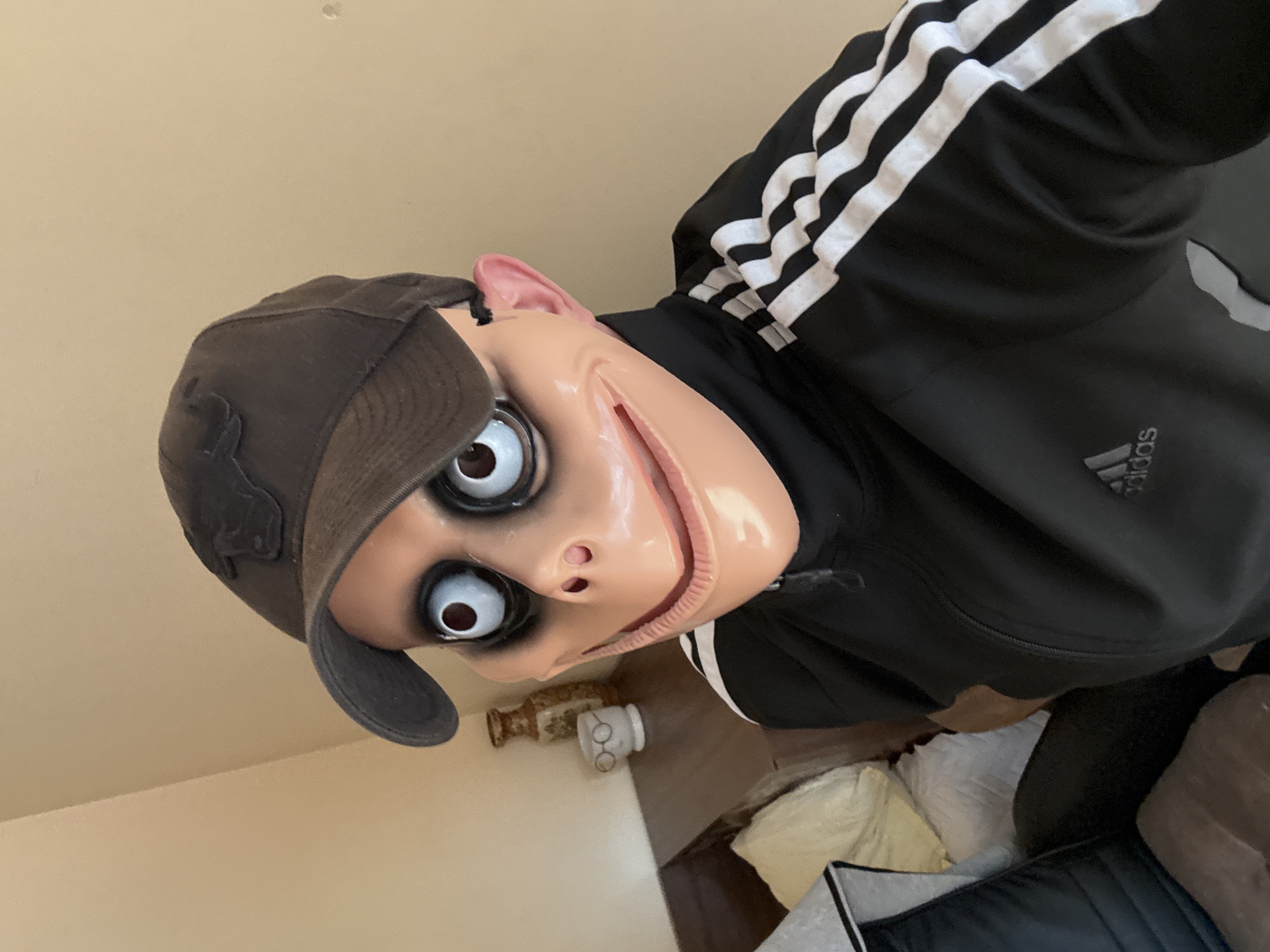 The image displays a tilted, eye-level indoor shot of a person wearing a creepy "Momo" mask. The mask features pale skin, large, unsettling white eyes with dark circles, and a wide, unnatural smile revealing gums. The person is also wearing a dark baseball cap, which sits on top of the mask's head. They are dressed in a black Adidas tracksuit top, clearly visible by the iconic white stripes on the sleeves and the "Adidas" logo on the right chest area.
The background primarily shows a plain light beige wall, indicating an indoor environment. Below the masked figure, on what appears to be a surface like a shelf or table, are a few objects: a rustic, possibly metallic bottle or vase, and a small white cylindrical item. Further down and to the right, indistinct soft furnishings, potentially pillows or cushions, are visible. The lighting suggests it is daytime. The scene captures a playful or spooky pose, likely for a costume or social media content. No specific visual elements within the image directly confirm the location as Varna, Bulgaria.